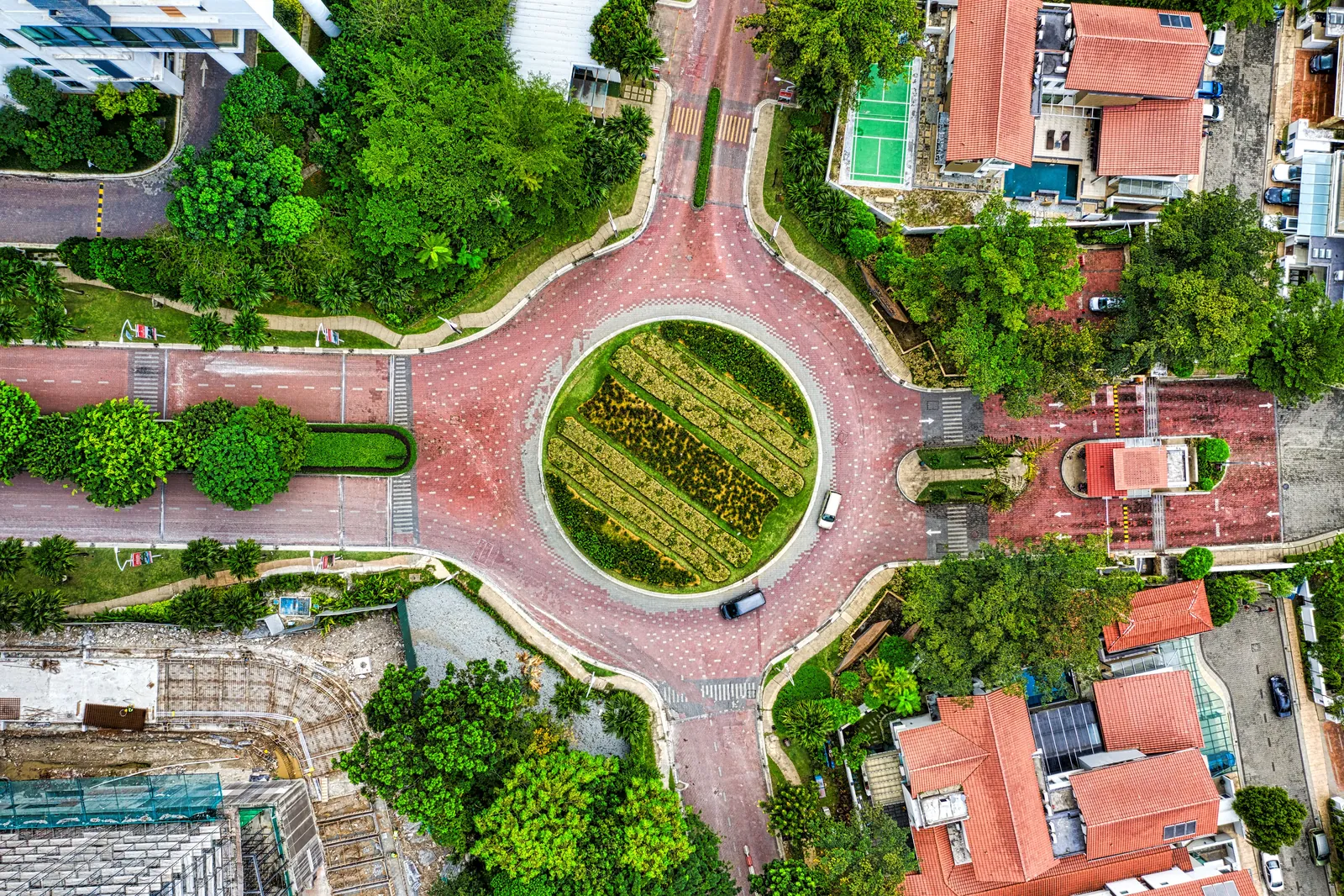 A roundabout paved in red with a round planted garden in the middle, seen from directly above, with tree-lined roads leading off, red-tiled house roofs, a small sports court and a building site at the lower left