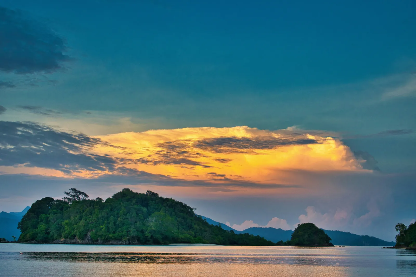 Wooded islands sit on a calm sea at dusk, under a bank of cloud lit orange by the setting sun