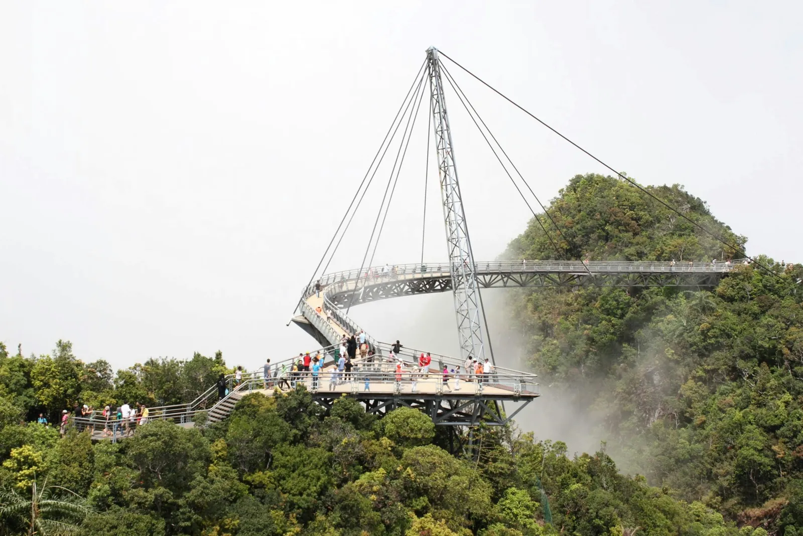 A curved steel footbridge hangs from a single tall pylon over a forested peak in the mist, with small groups of visitors walking along it