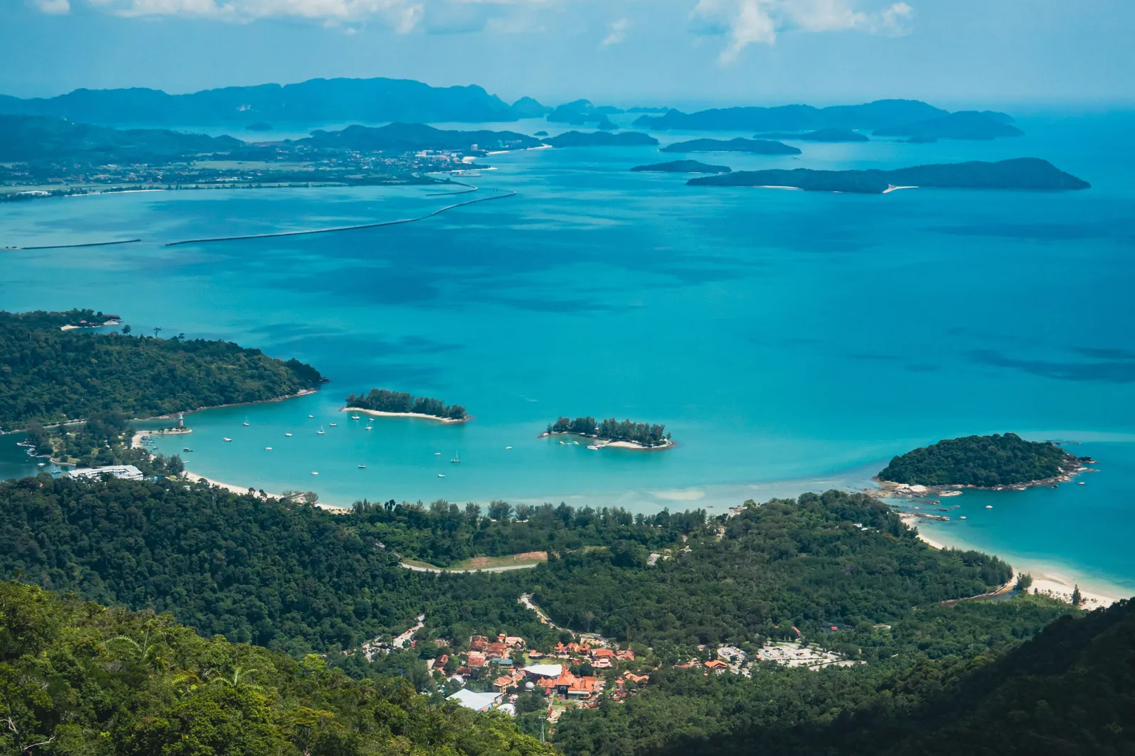 Rainforest falls away to a turquoise bay dotted with small wooded islands, with red-roofed buildings among the trees and more islands on the horizon