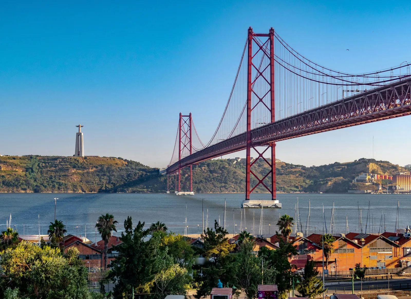 A long red suspension bridge over a wide river, with a tall statue on the hill across the water and palm trees and red roofs in the foreground