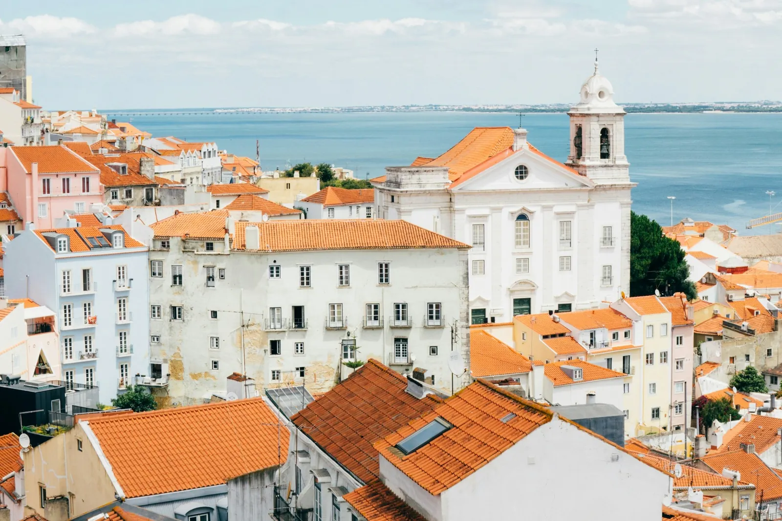 Orange-tiled roofs and white and pink houses packed down a hillside to a white church with a bell tower, with the wide river behind