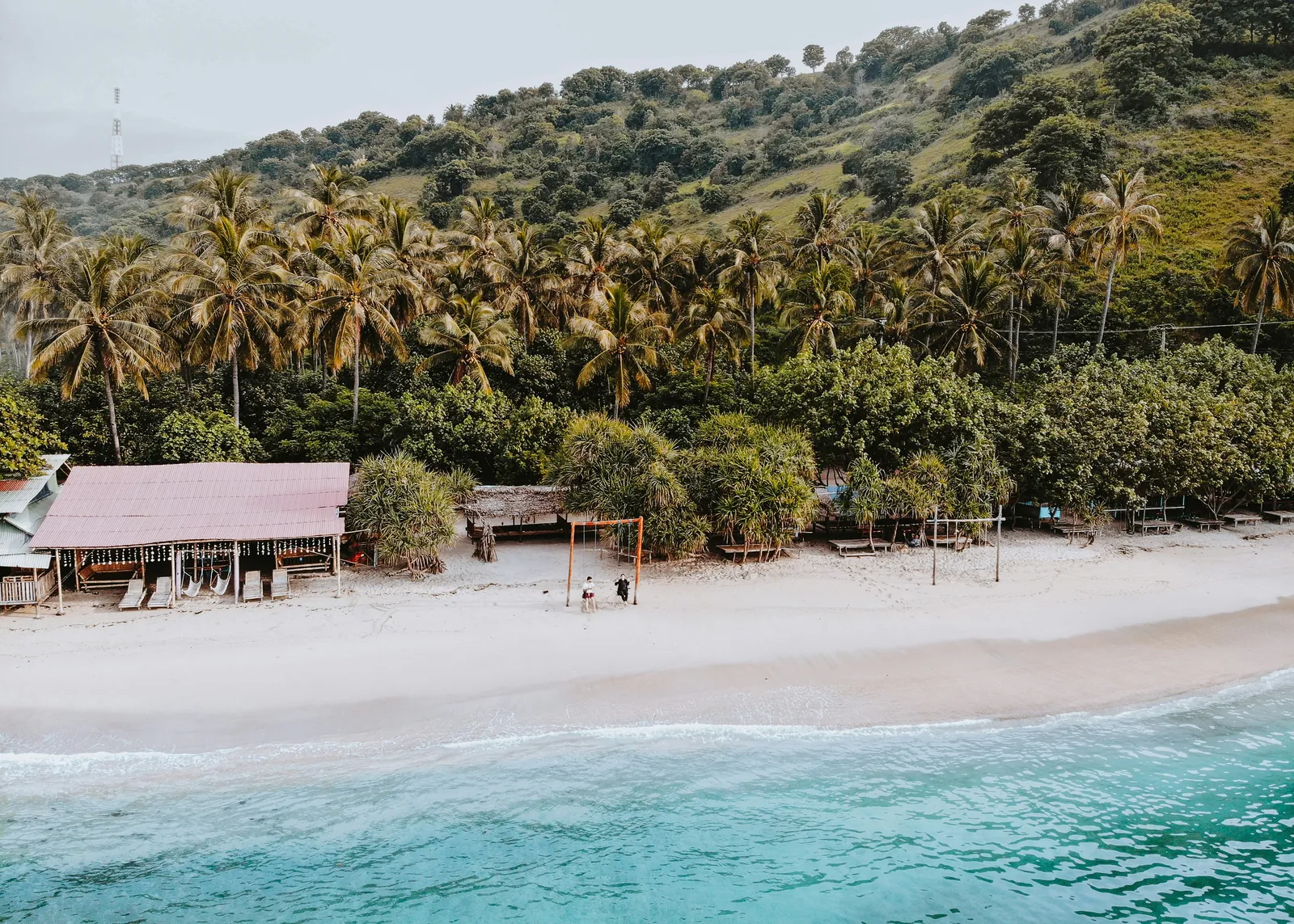 A white sand beach with a long open-sided shelter, sun loungers and a swing, palm and pandanus trees and a green hillside behind, seen from above