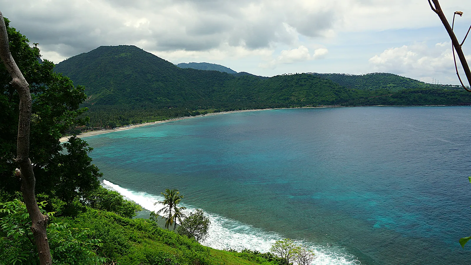 A turquoise bay with a curve of sandy beach below steep green hills, under a cloudy sky