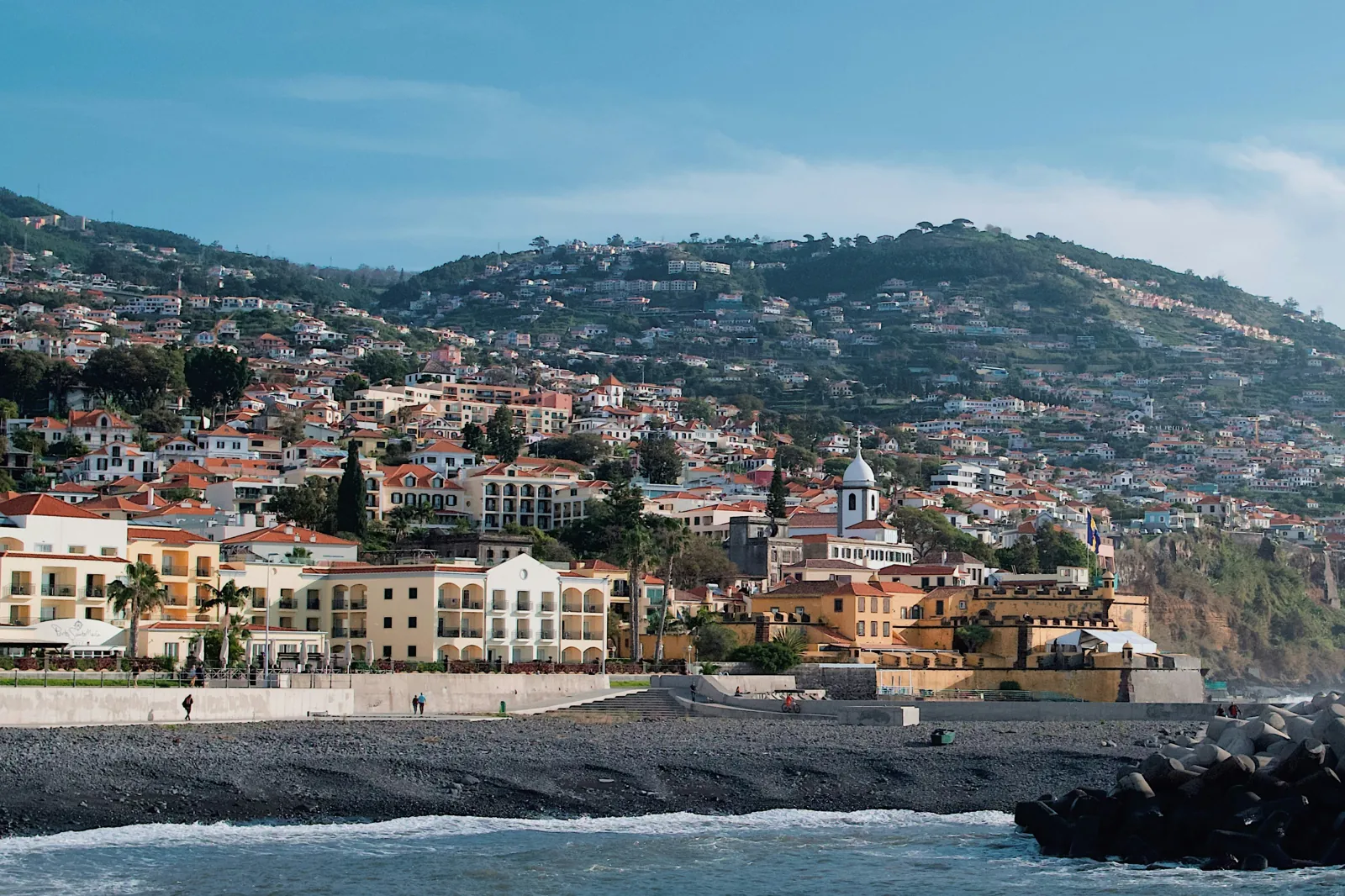 Funchal’s seafront, with a stony beach in front and yellow and white buildings with red roofs climbing steep green hills behind