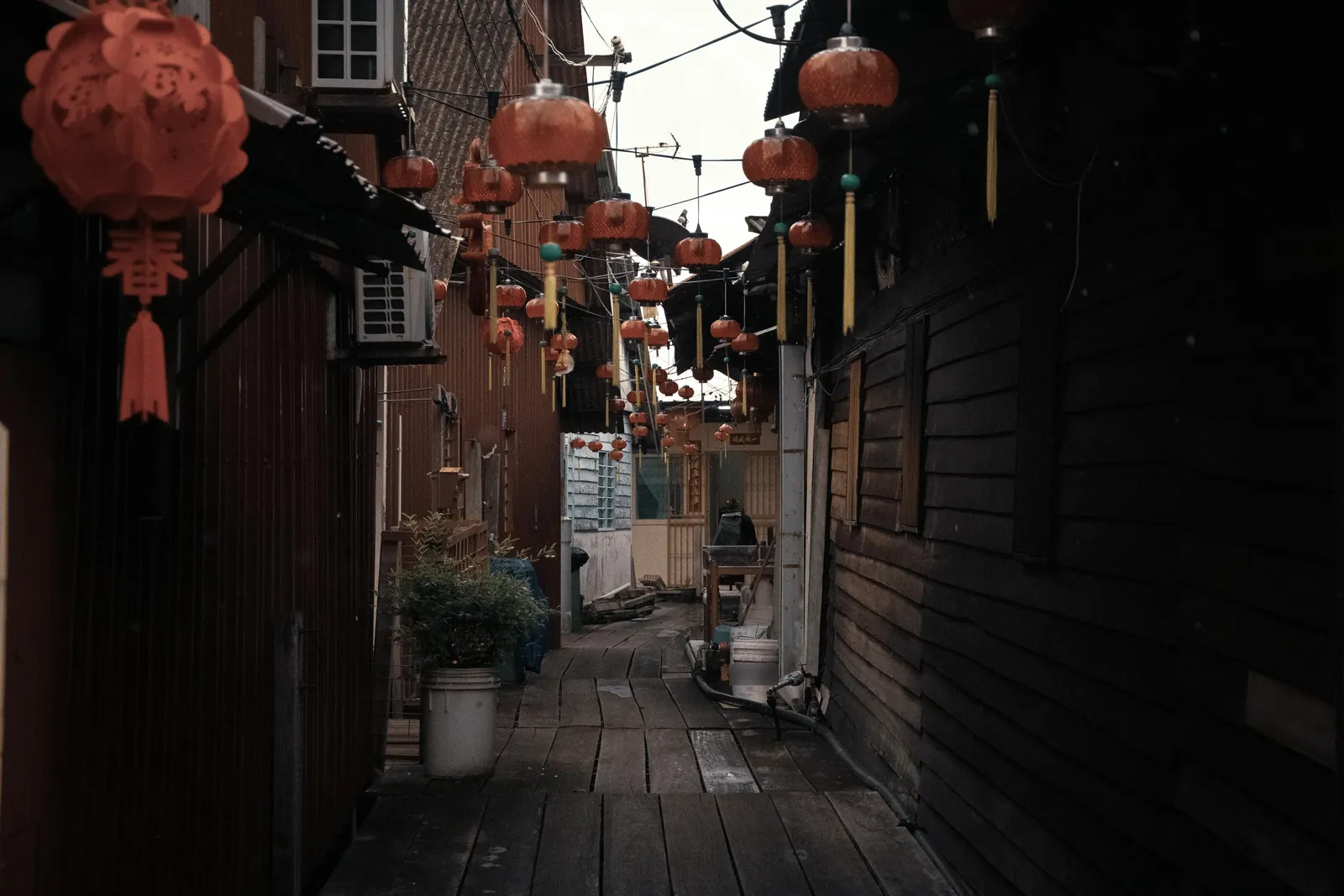 Narrow wooden boardwalk between dark timber houses, with red paper lanterns strung overhead and potted plants along the side