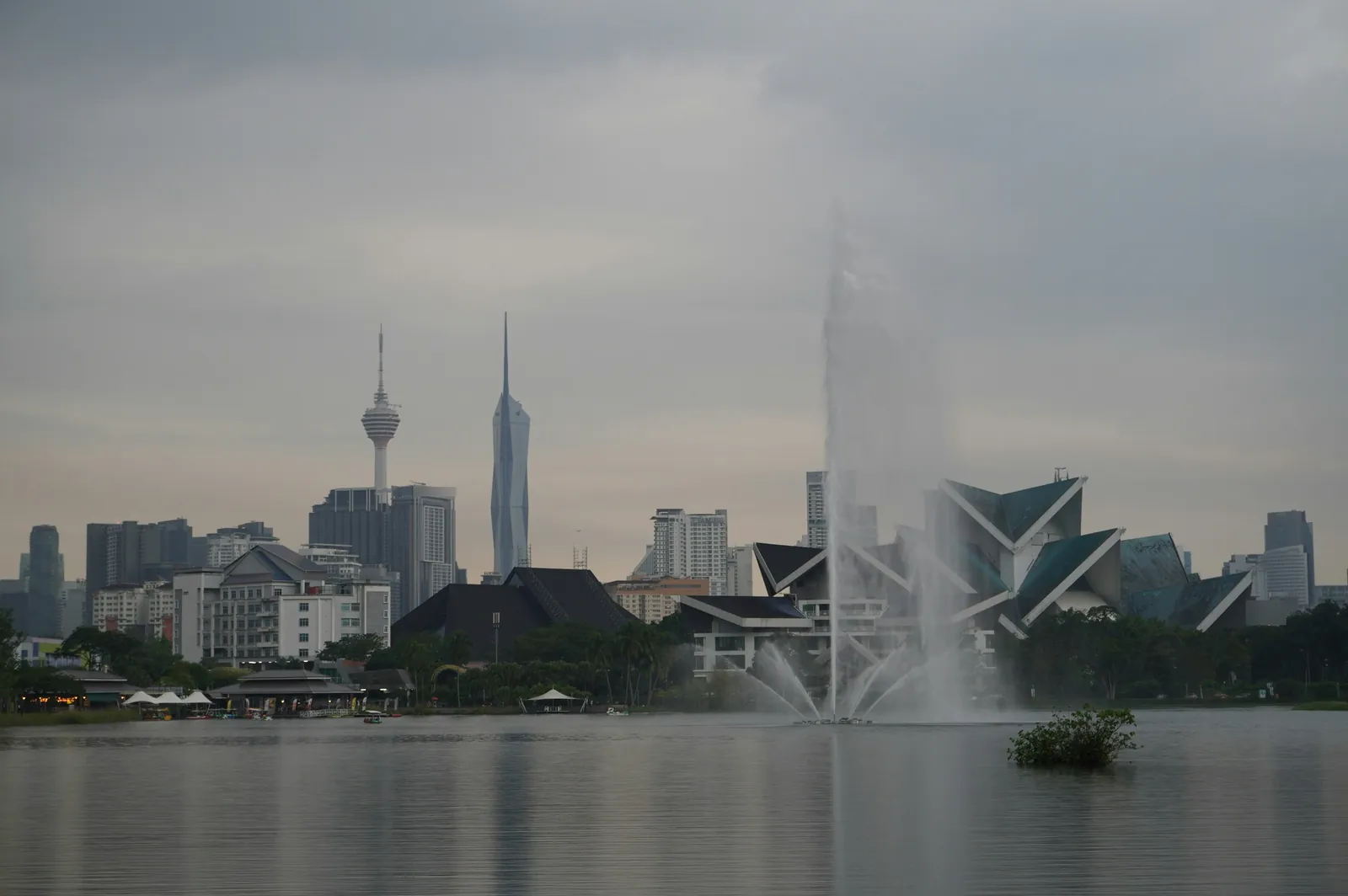 Kuala Lumpur skyline with the KL Tower and Merdeka 118 rising behind a park lake with a tall fountain and a folded green roof on the right
