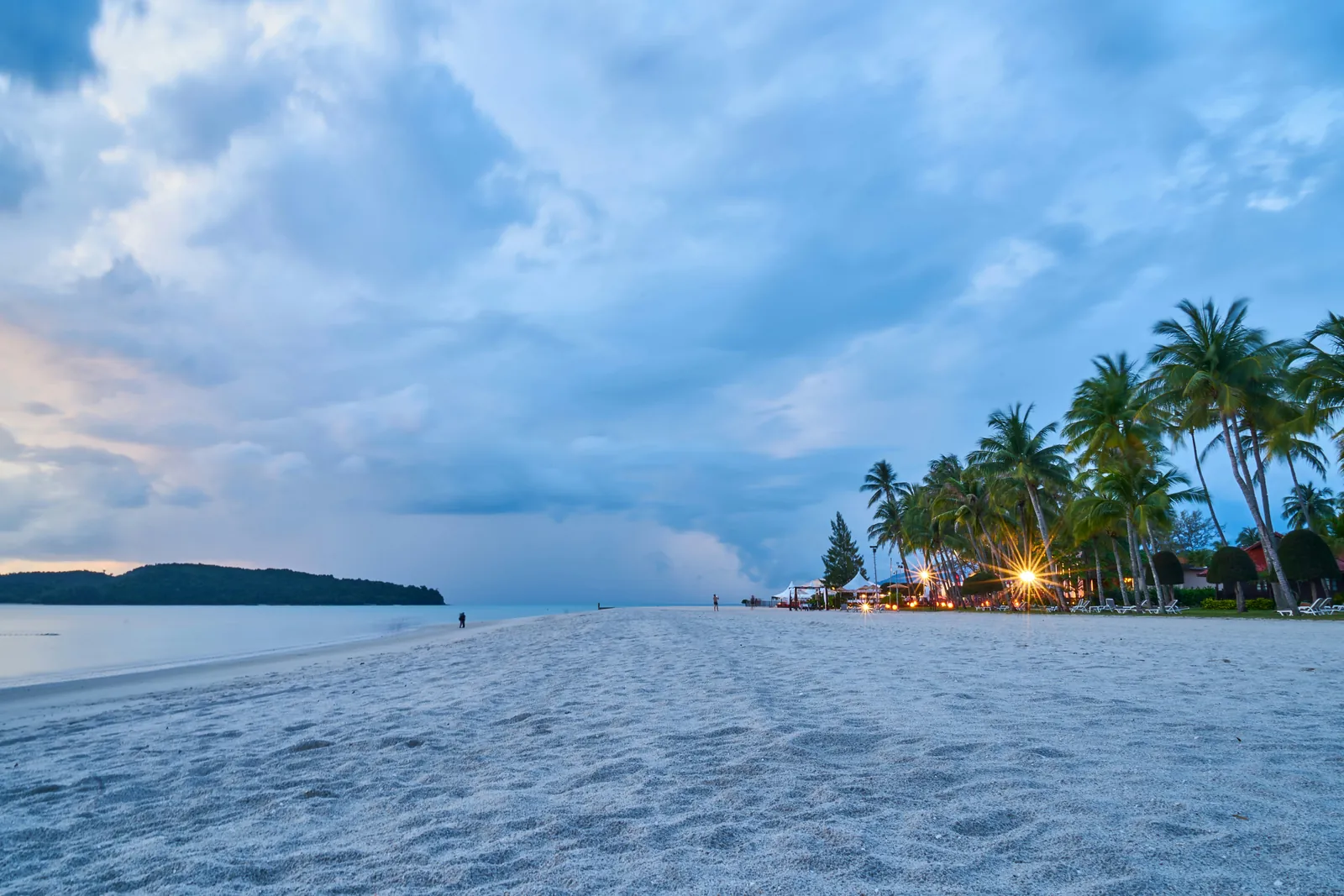 Wide white beach at dusk under a cloudy blue sky, with coconut palms and warm lights on the right and a wooded island across the water