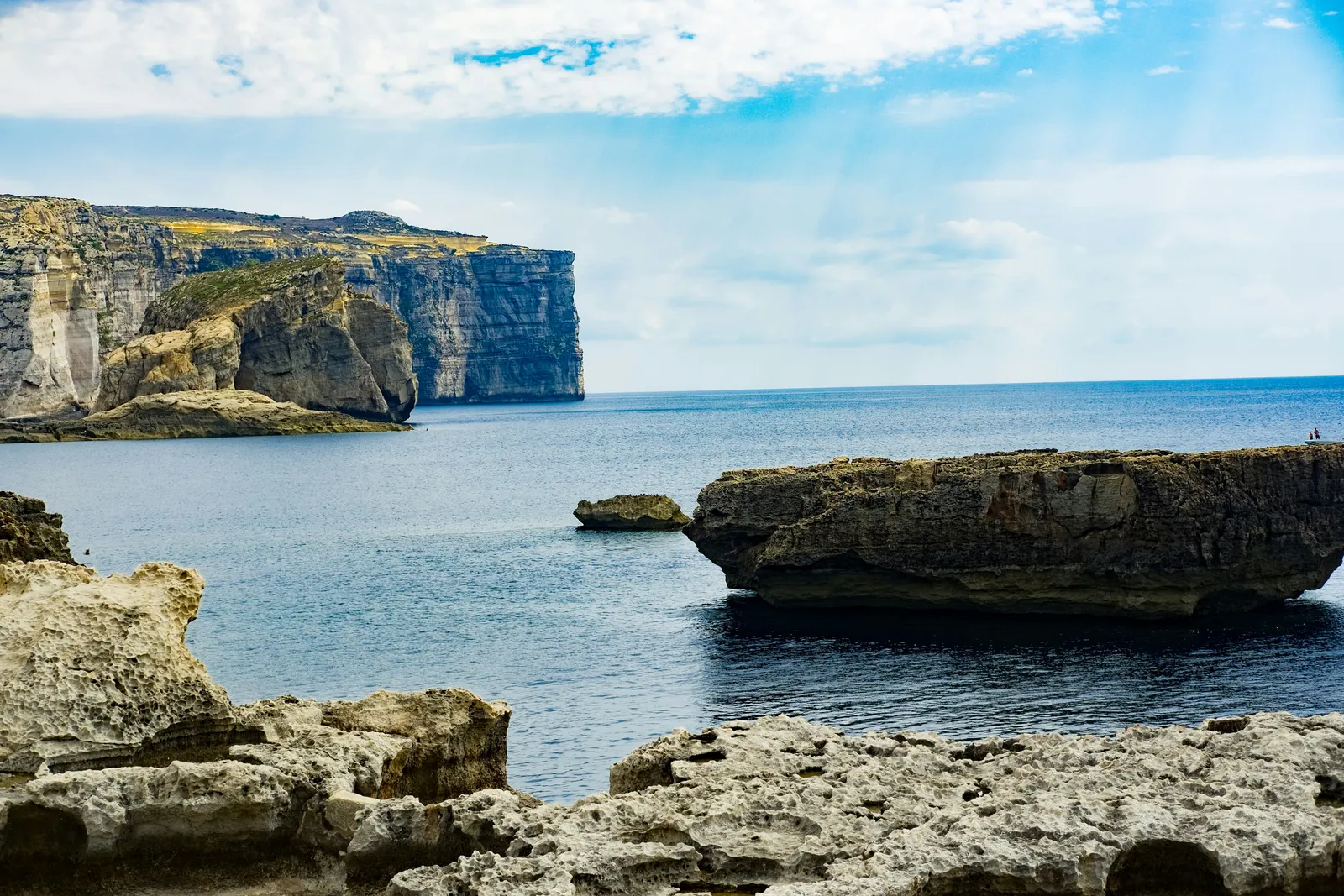 Limestone rocks in the foreground and Fungus Rock below high sea cliffs at Dwejra Bay on Gozo, under a bright partly cloudy sky