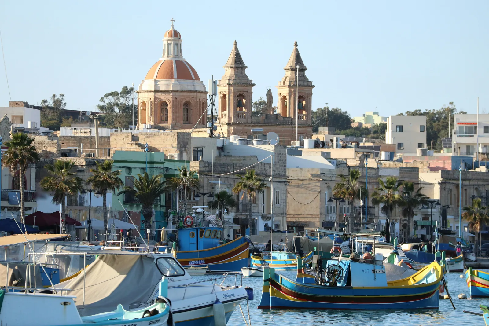 Brightly painted luzzu fishing boats in Marsaxlokk harbour, with palm trees, waterfront houses and the domed parish church behind