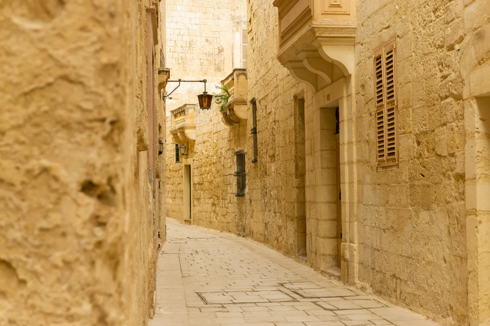 A narrow curving lane between honey-coloured limestone walls in Mdina, with an iron lantern, wooden shutters and small stone balconies