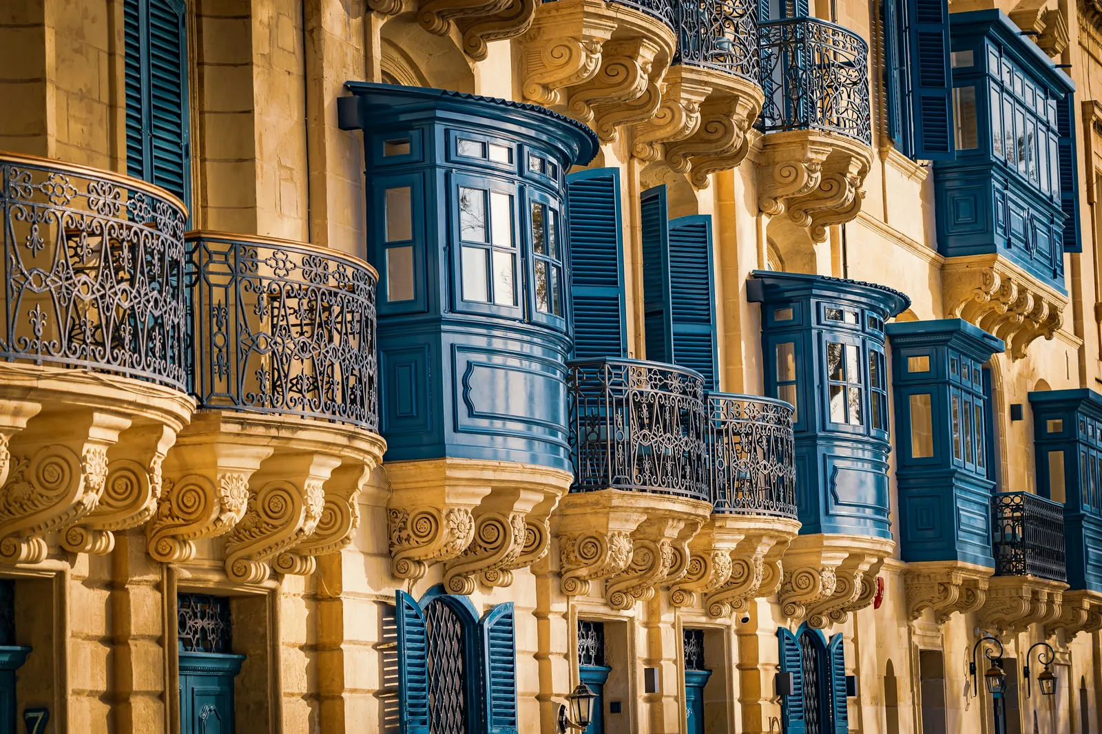 A row of honey-coloured limestone houses in Valletta with blue enclosed wooden balconies, wrought-iron balconies and carved stone brackets