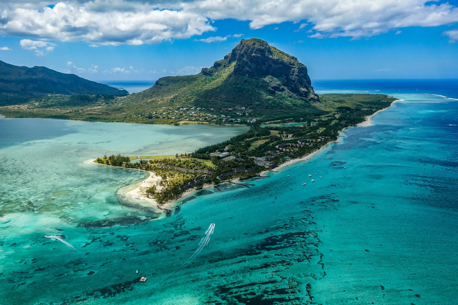 A tall dark mountain with sheer cliffs rising above a low green peninsula fringed with white beaches, palm groves and low hotel roofs, surrounded by a turquoise lagoon and reef, seen from the air