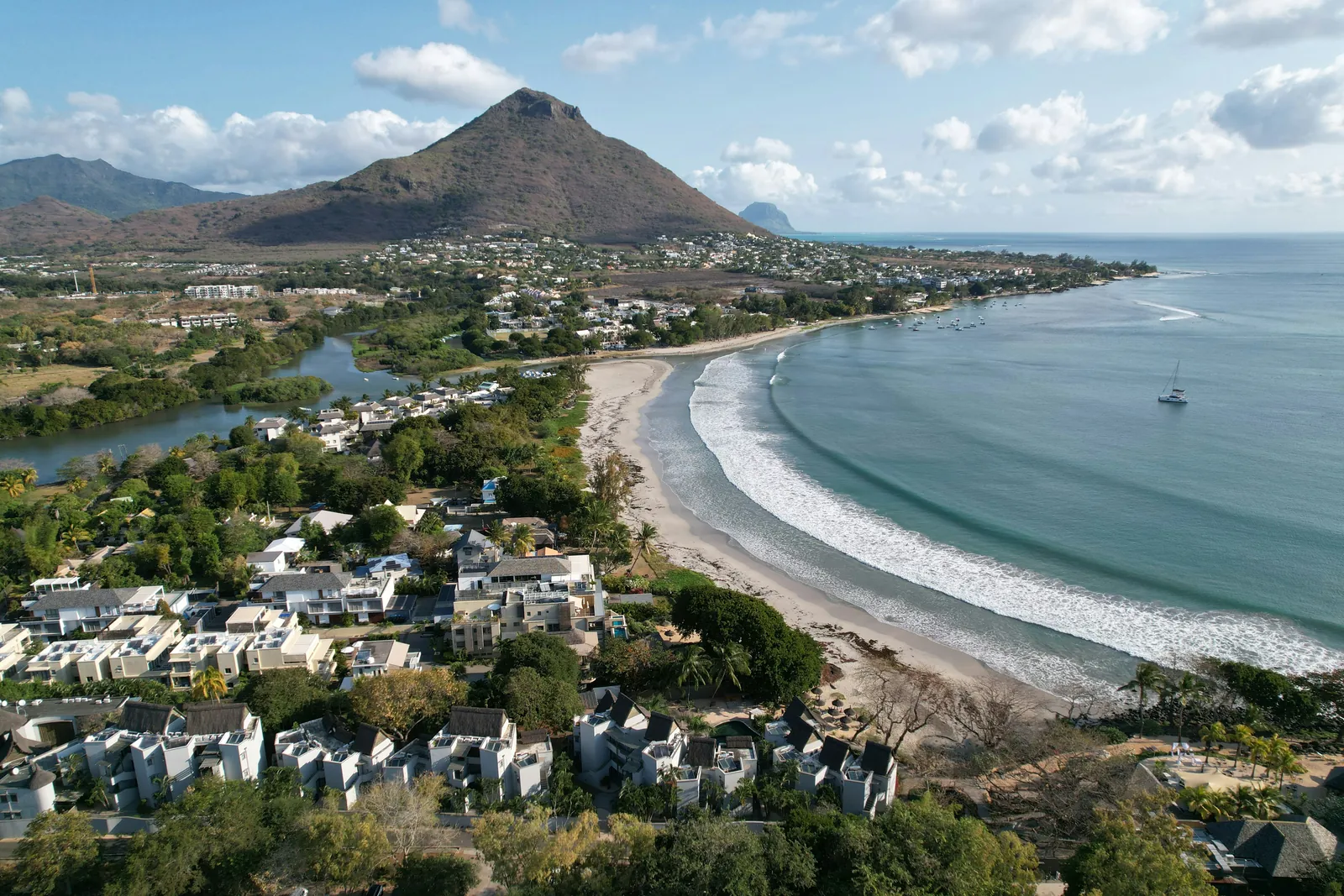 A curved sandy beach and wide bay with rolling surf, a river mouth and low white houses among trees, and a pointed brown mountain behind, seen from a drone