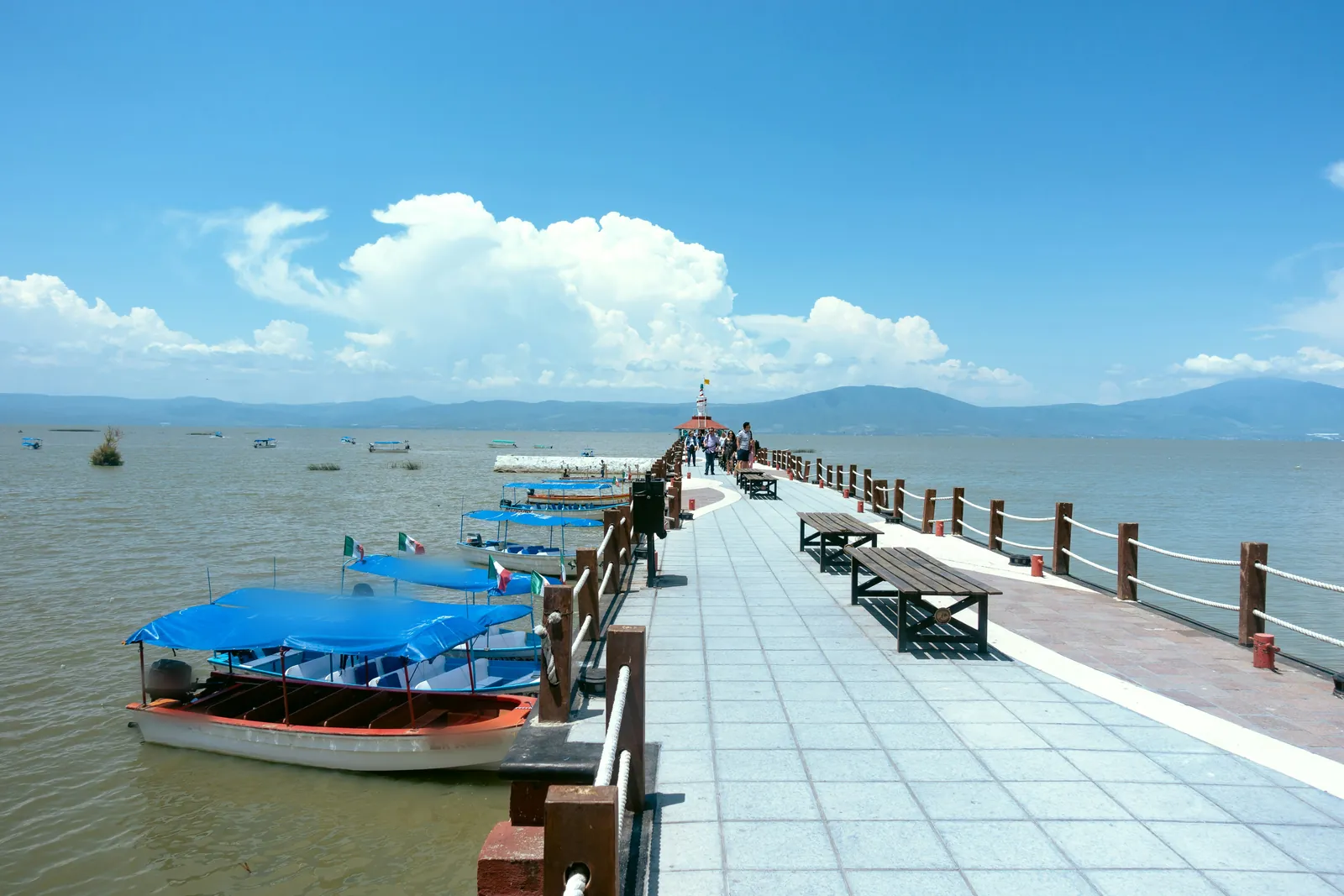 A long tiled pier with wooden benches and a rope rail running out into a wide lake, with canopied boats flying Mexican flags moored alongside and hills on the far shore