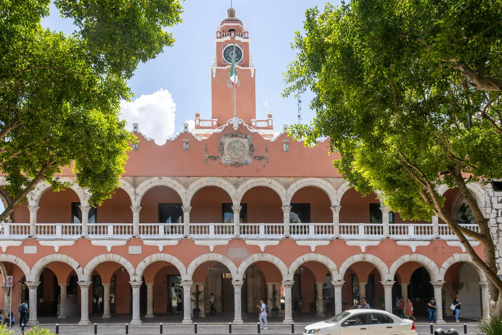 A two-storey colonial building painted coral pink with a long white arcade of arches on both floors and a clock tower flying the Mexican flag, framed by two leafy trees, with people walking under the arches