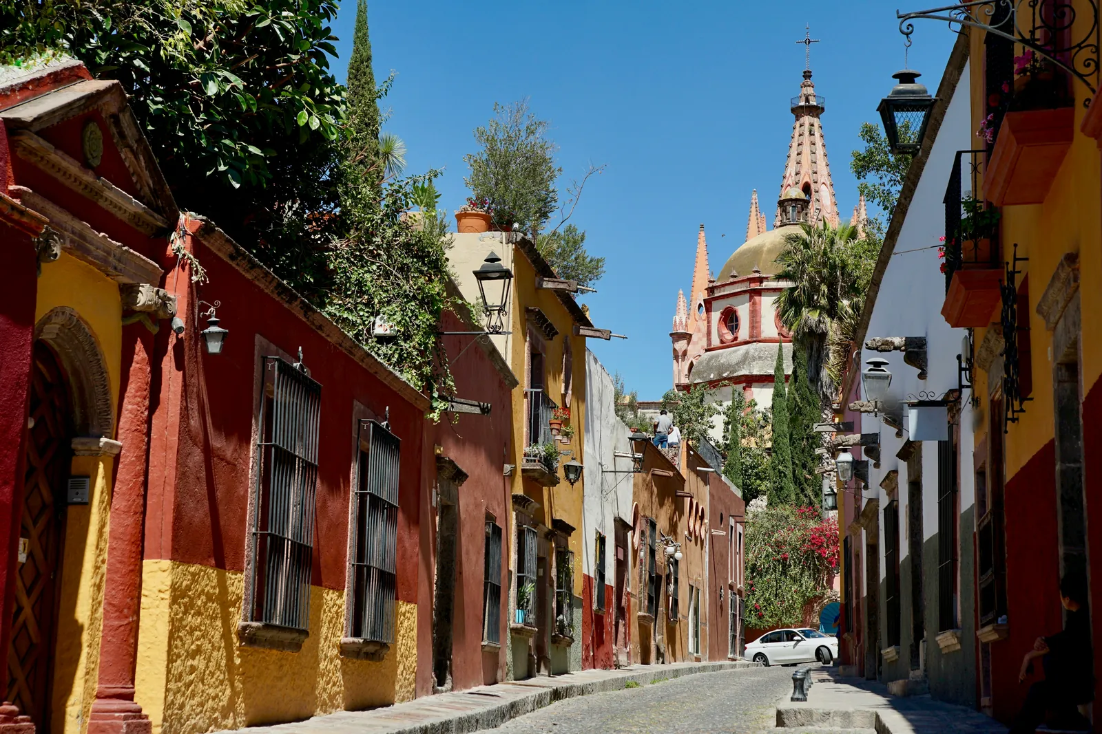 A sunny cobbled street of red, ochre and yellow colonial houses with iron window grilles and lanterns, leading towards the pink spires of a large church