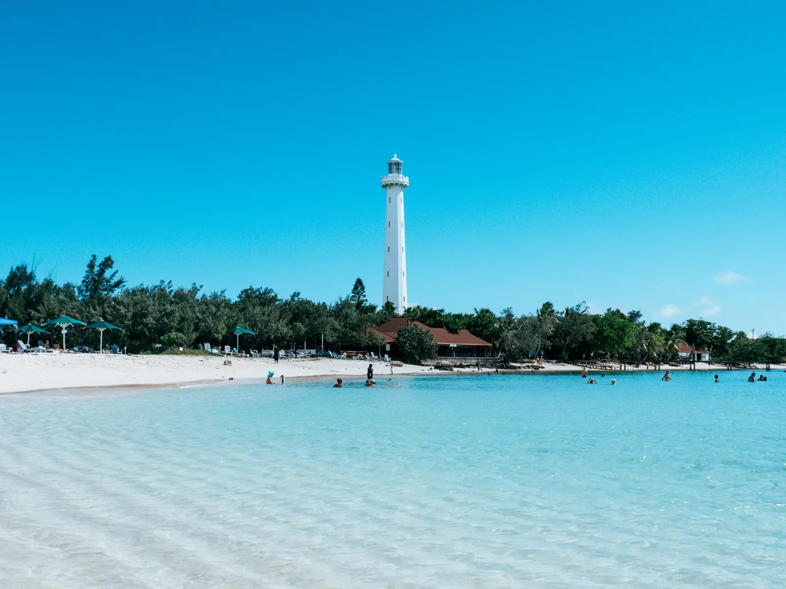 A slim white lighthouse rises above trees behind a white beach and a pale turquoise lagoon, with small figures swimming in the shallows
