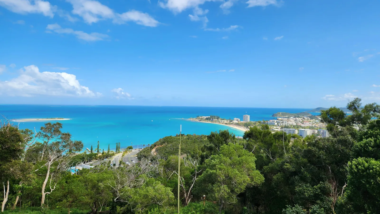 Bush on a hillside slopes down to a bay with a curved beach and apartment buildings, with a turquoise lagoon beyond