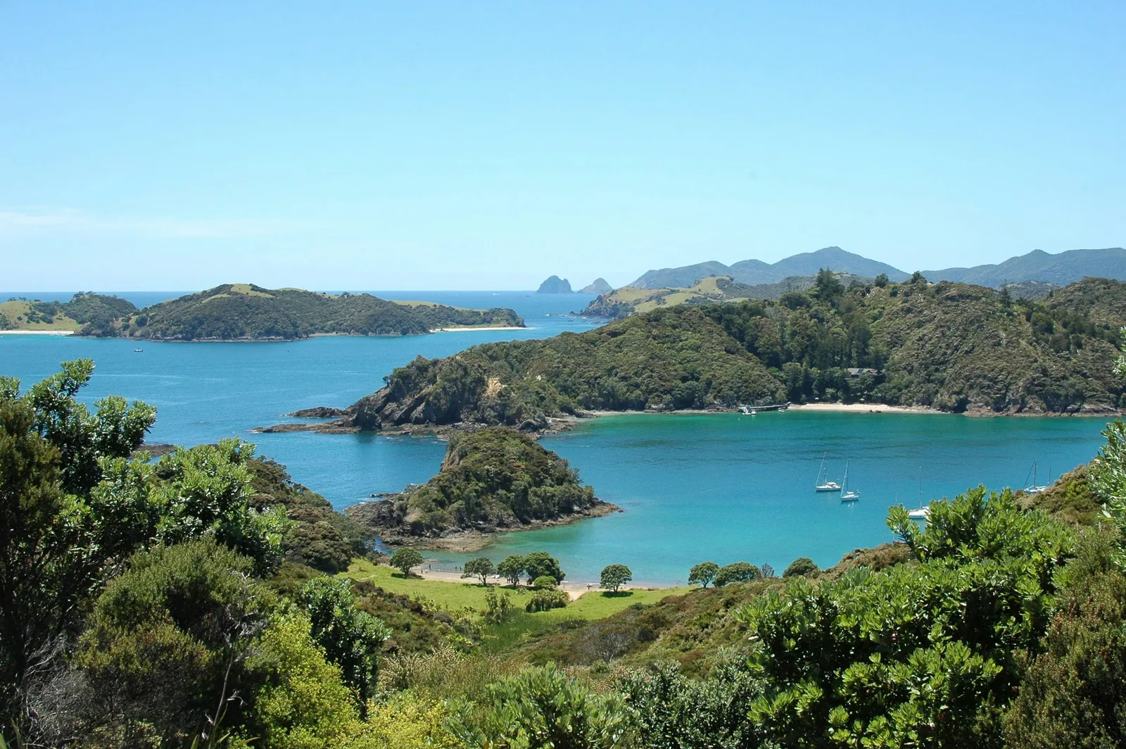 Bush-covered islands and headlands around a turquoise bay with a few yachts at anchor, in the Bay of Islands