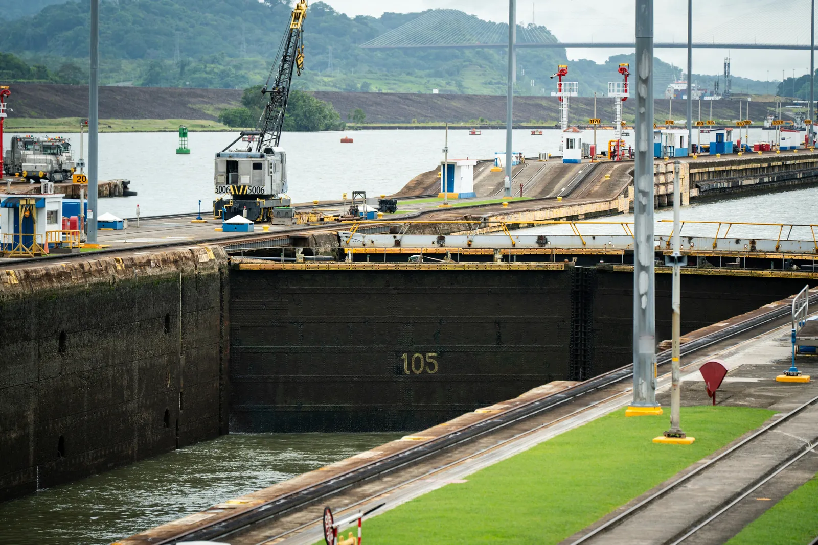 A deep concrete lock chamber with a tall dark steel gate numbered 105, a crane and towing locomotive on the lock wall, and a cable-stayed bridge over green hills behind