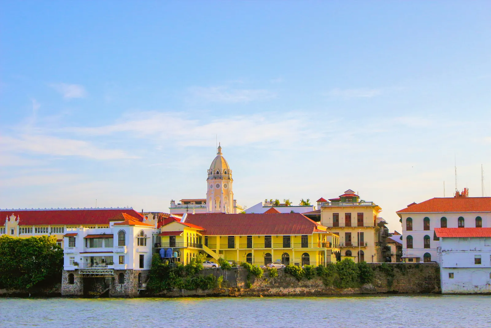 A waterfront row of white, yellow and cream colonial buildings with red tiled roofs on an old sea wall, a domed church tower rising behind, in warm late light