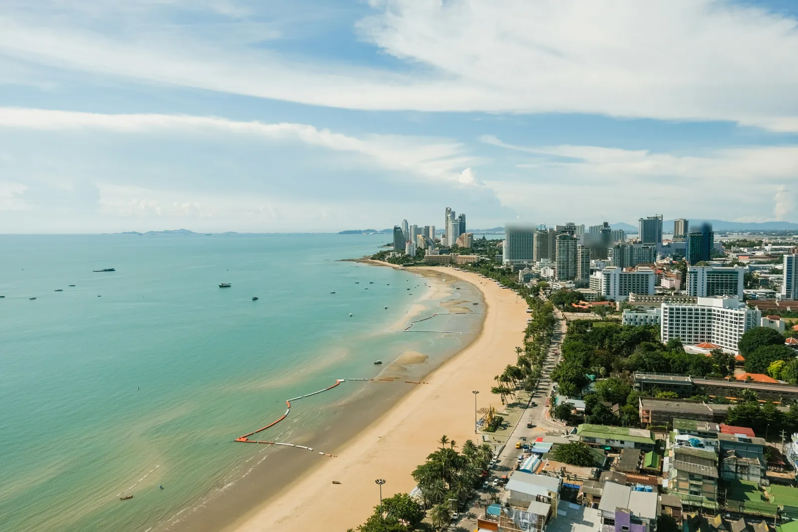 A high view along the curve of Pattaya Beach at low tide, with a palm-lined beach road, hotels and flats behind, and small boats in a calm green bay