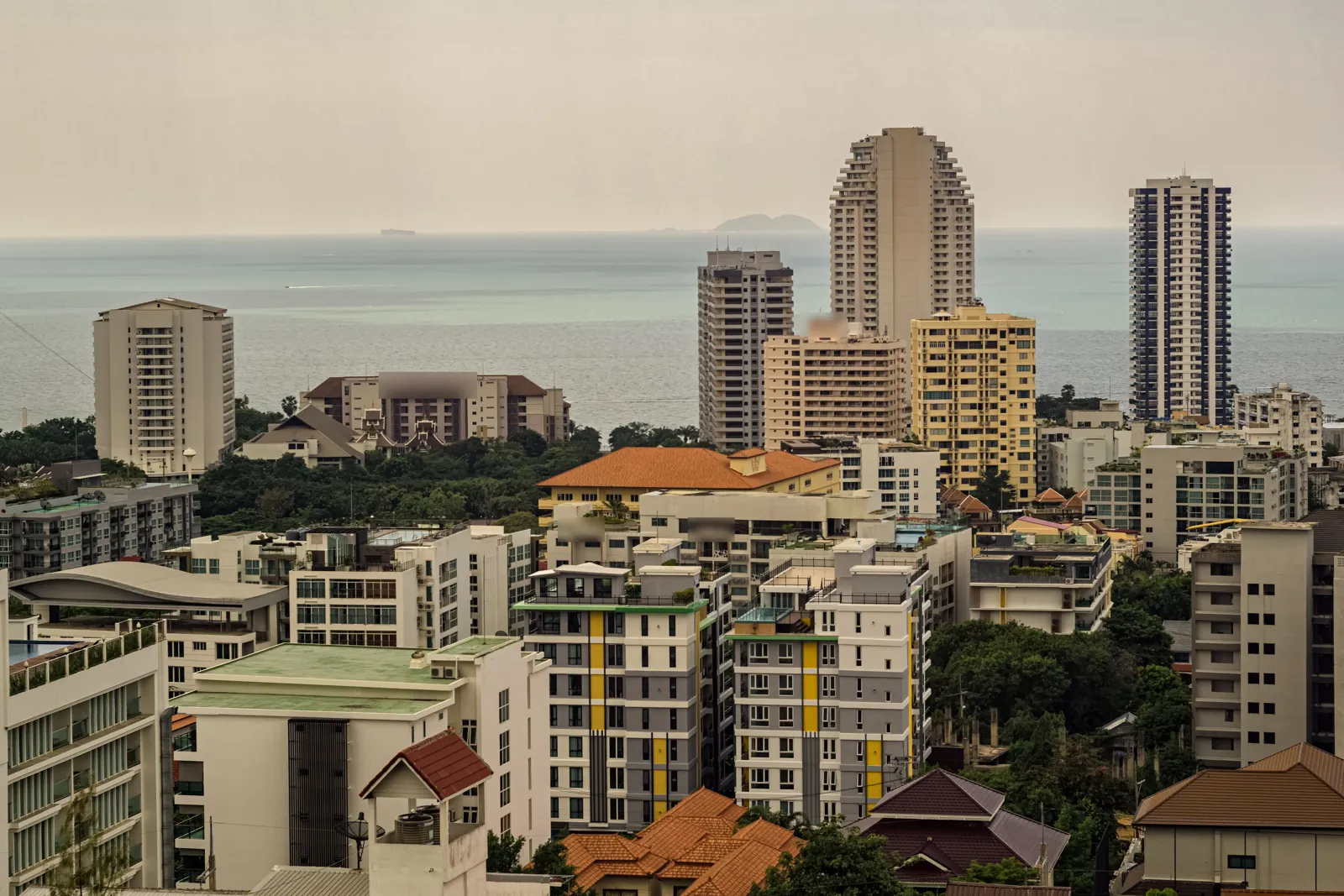 Condominium and hotel towers and low-rise buildings among trees, with a hazy sea and a small island behind