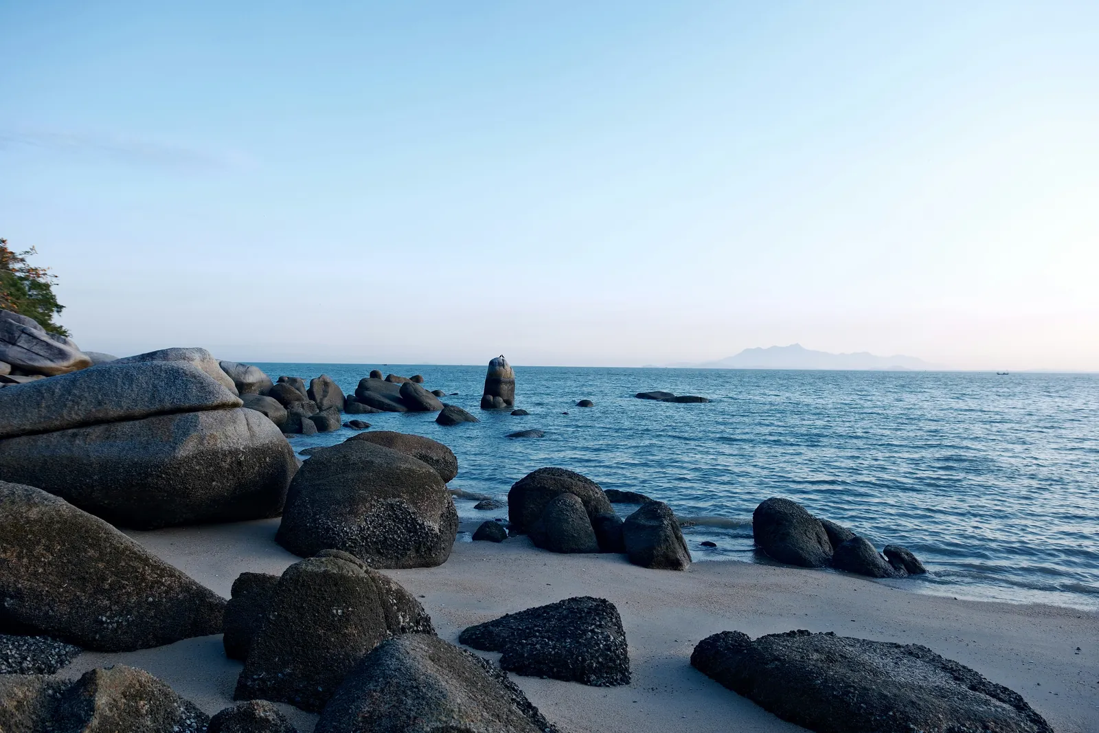 Large smooth dark boulders scattered along a pale sandy beach and out into a calm blue sea, with a hazy coastline on the horizon under a clear sky