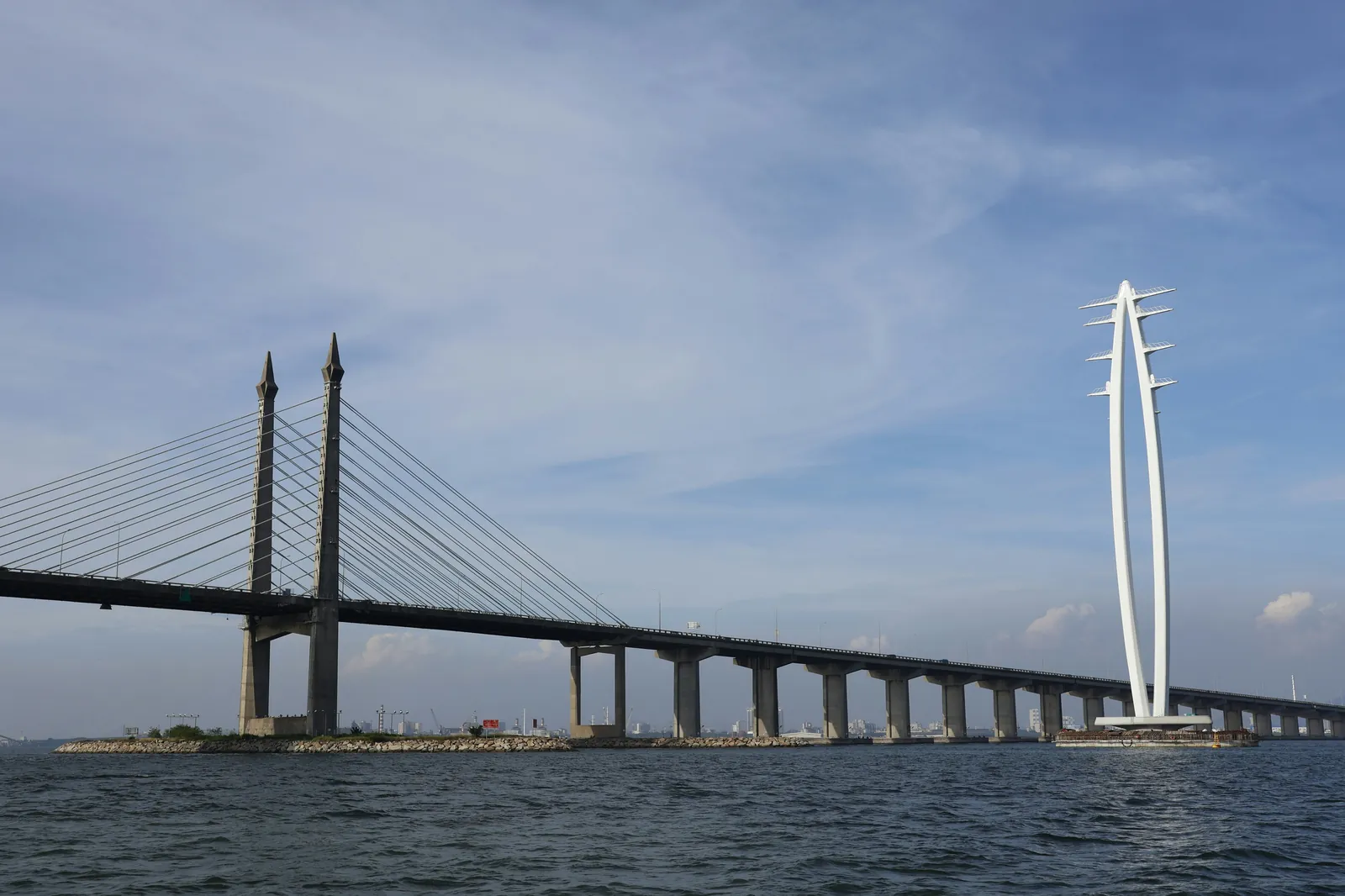 A long road bridge on concrete piers crossing open water, with two tall pointed towers holding fans of cables, and a tall white arch-shaped sculpture on a small island beside it, under a blue sky with light cloud
