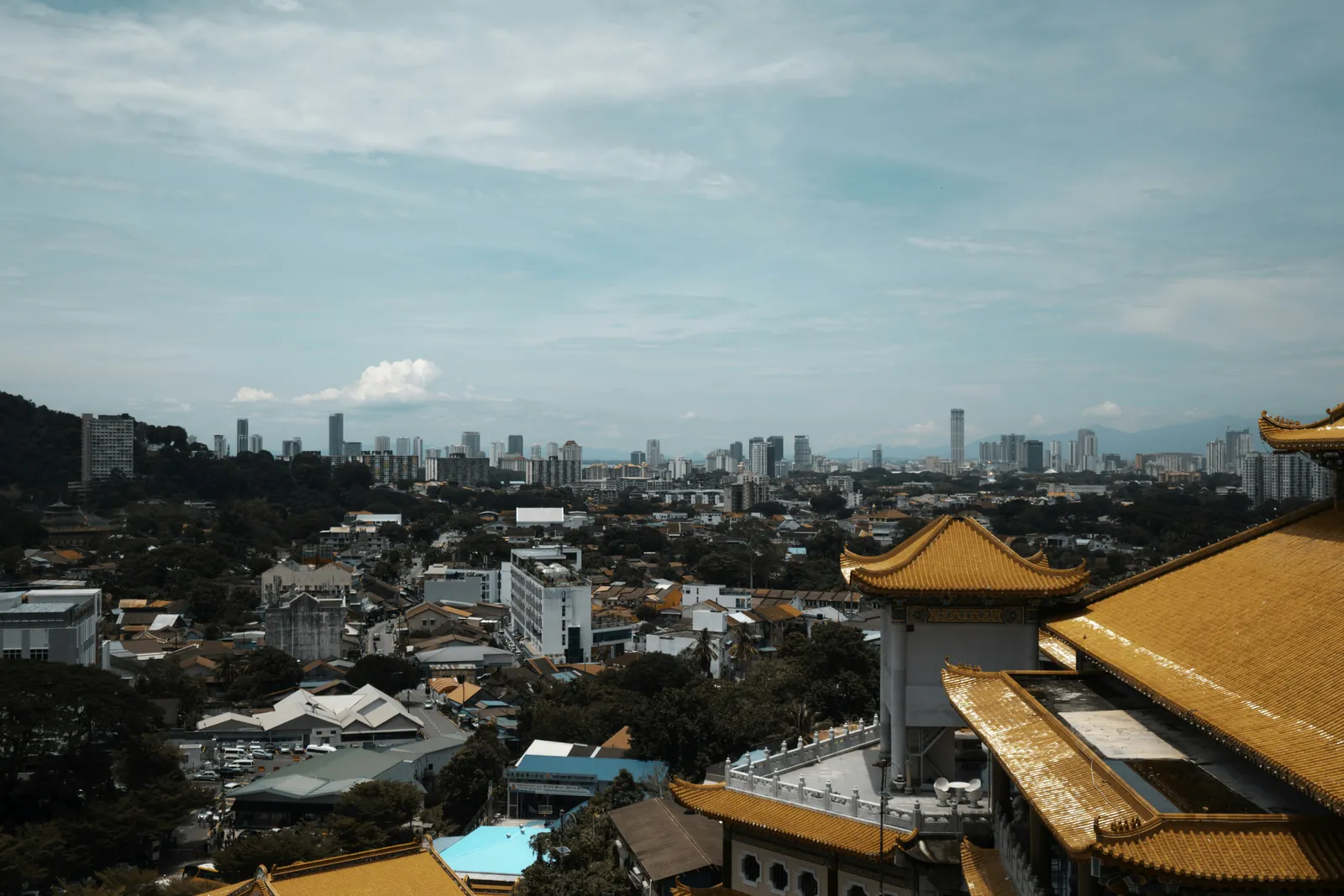 Curved golden-yellow tiled temple roofs in the foreground at the right, looking out over low houses and trees to a line of high-rise towers on the horizon under a pale blue sky