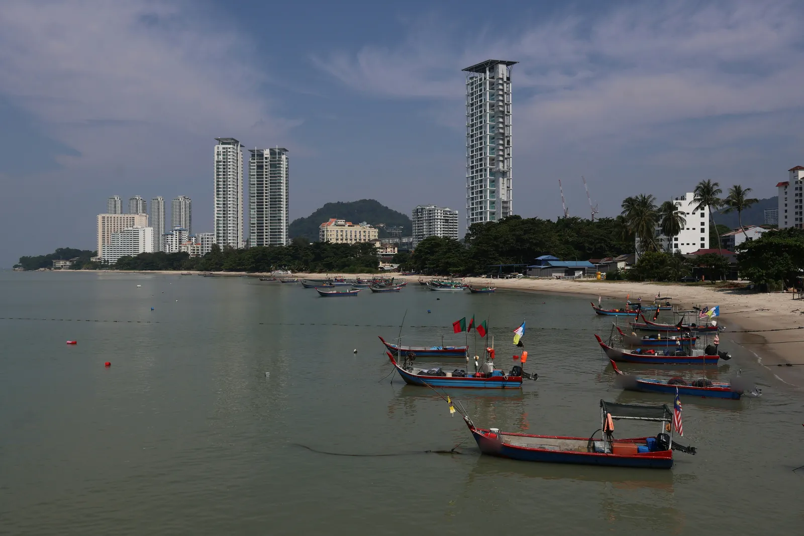 Small blue and red wooden fishing boats with flags anchored in calm shallow water off a sandy beach, with white condominium towers, palm trees and a wooded hill behind under a blue sky