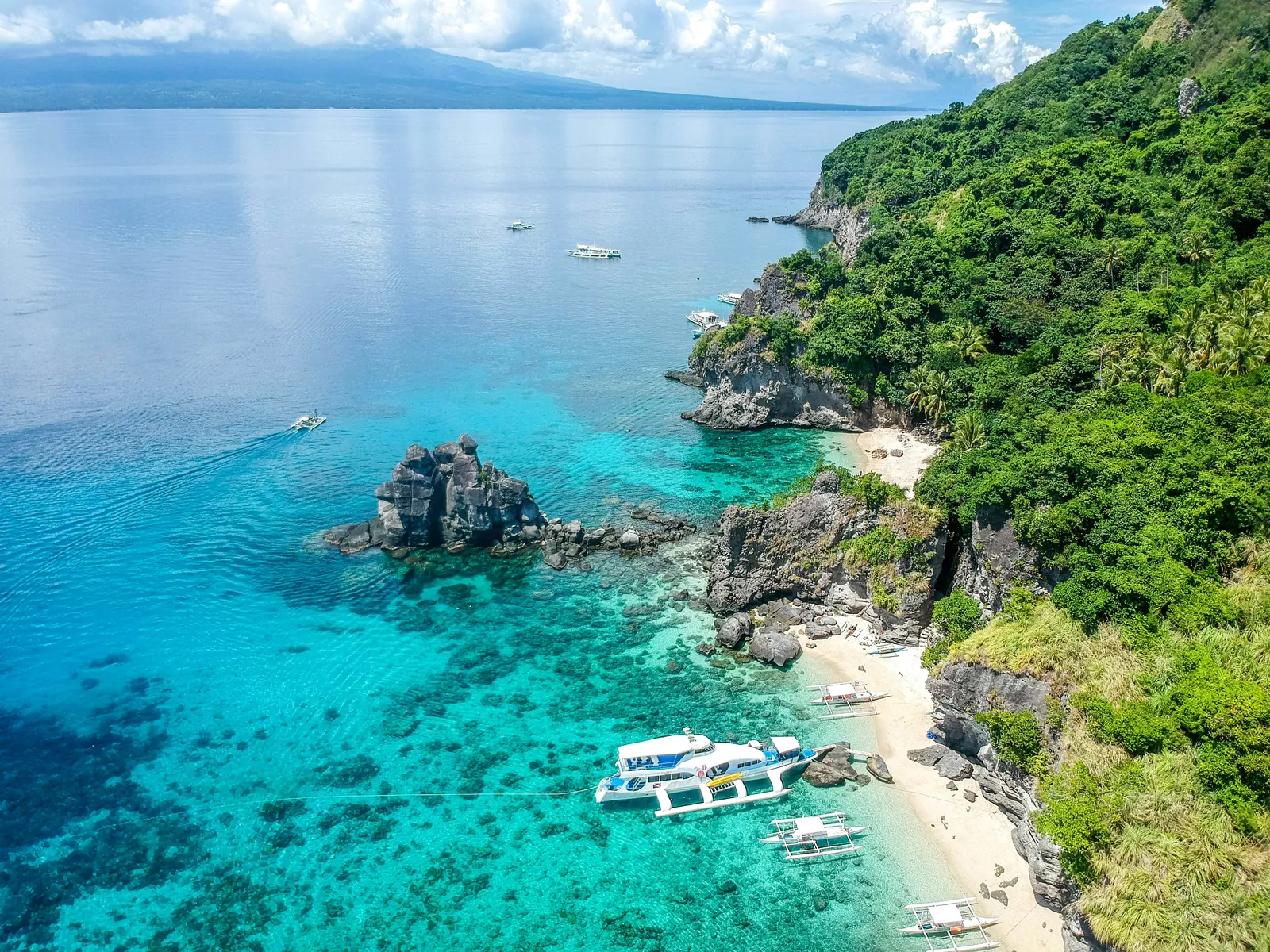 Aerial view of clear turquoise water over coral, dark rocks and a small white beach below a green hillside, with outrigger boats pulled in