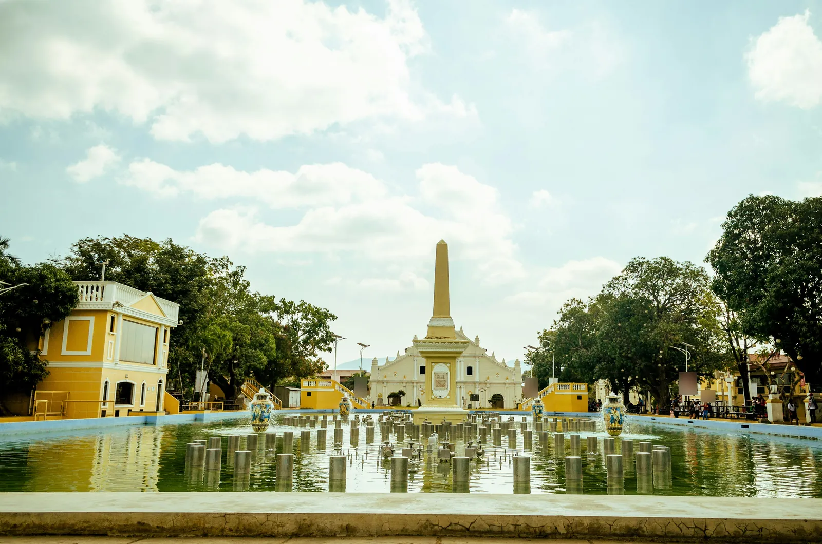 Long fountain pool leading to a tall obelisk and the white facade of an old cathedral, with yellow colonial buildings and trees on each side