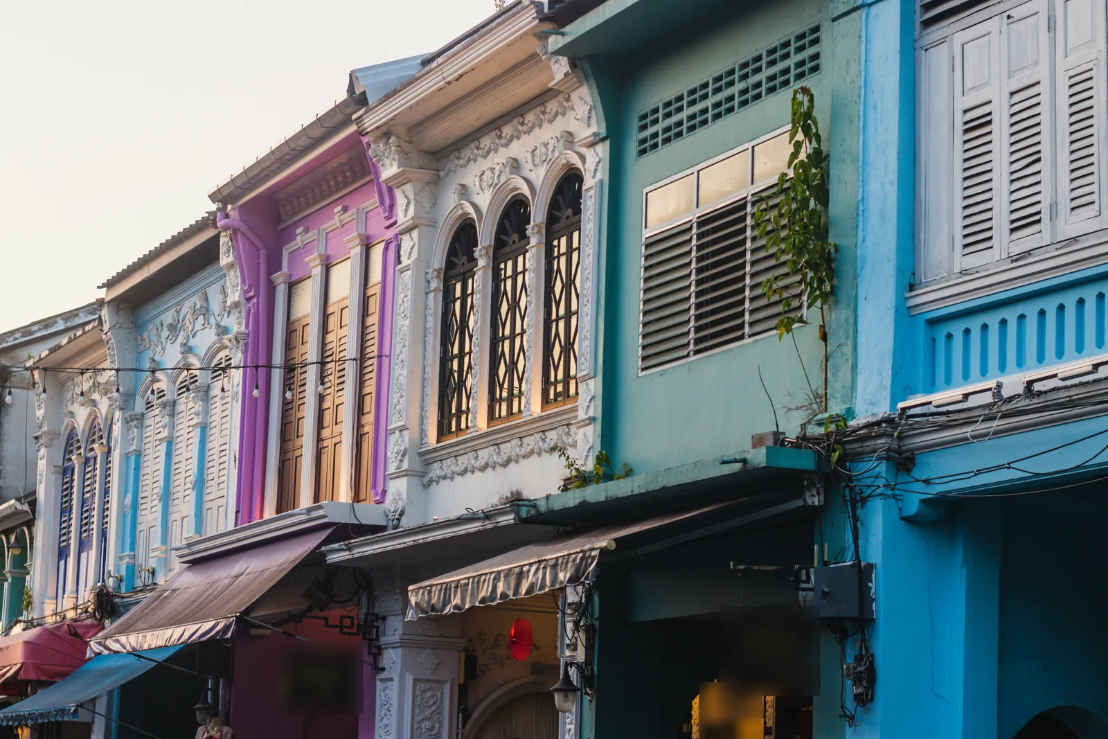 The upper storeys of a row of old shophouses painted pink, white, mint and blue, with arched windows, wooden shutters and awnings, in late afternoon light