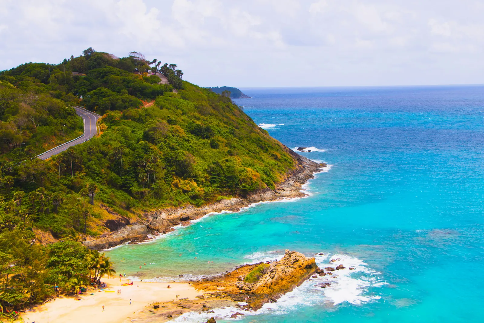 A green headland with a winding road above a small sandy beach and a turquoise sea, with another headland on the horizon