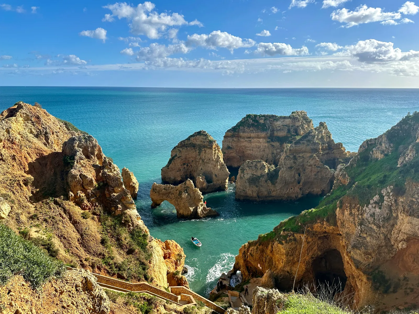 Golden sandstone cliffs, sea stacks and a rock arch rising from turquoise water, with a small boat in the cove and steps down the cliff