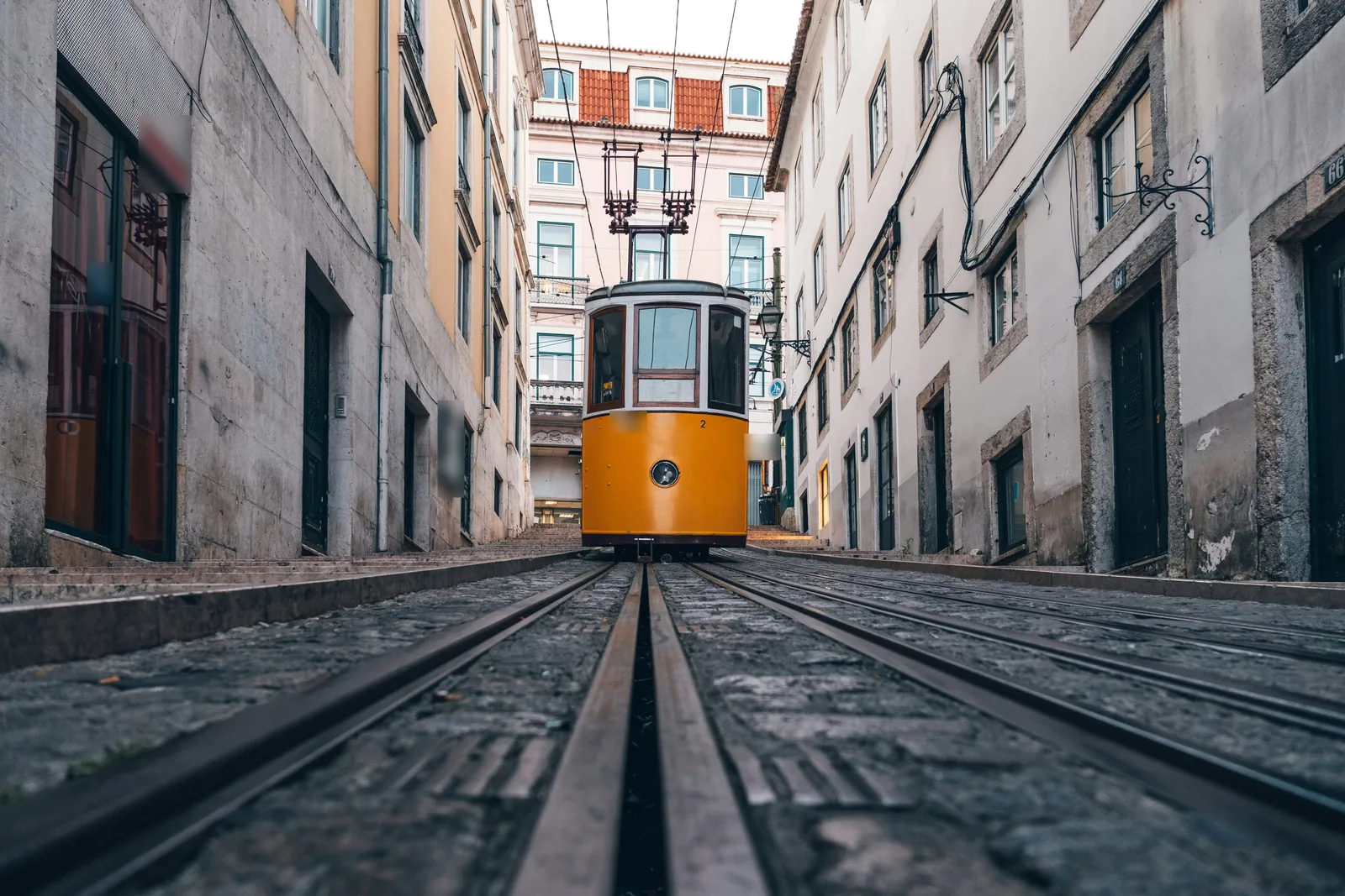 Low view along tram rails up a steep cobbled street to a small yellow tram car between pale old buildings