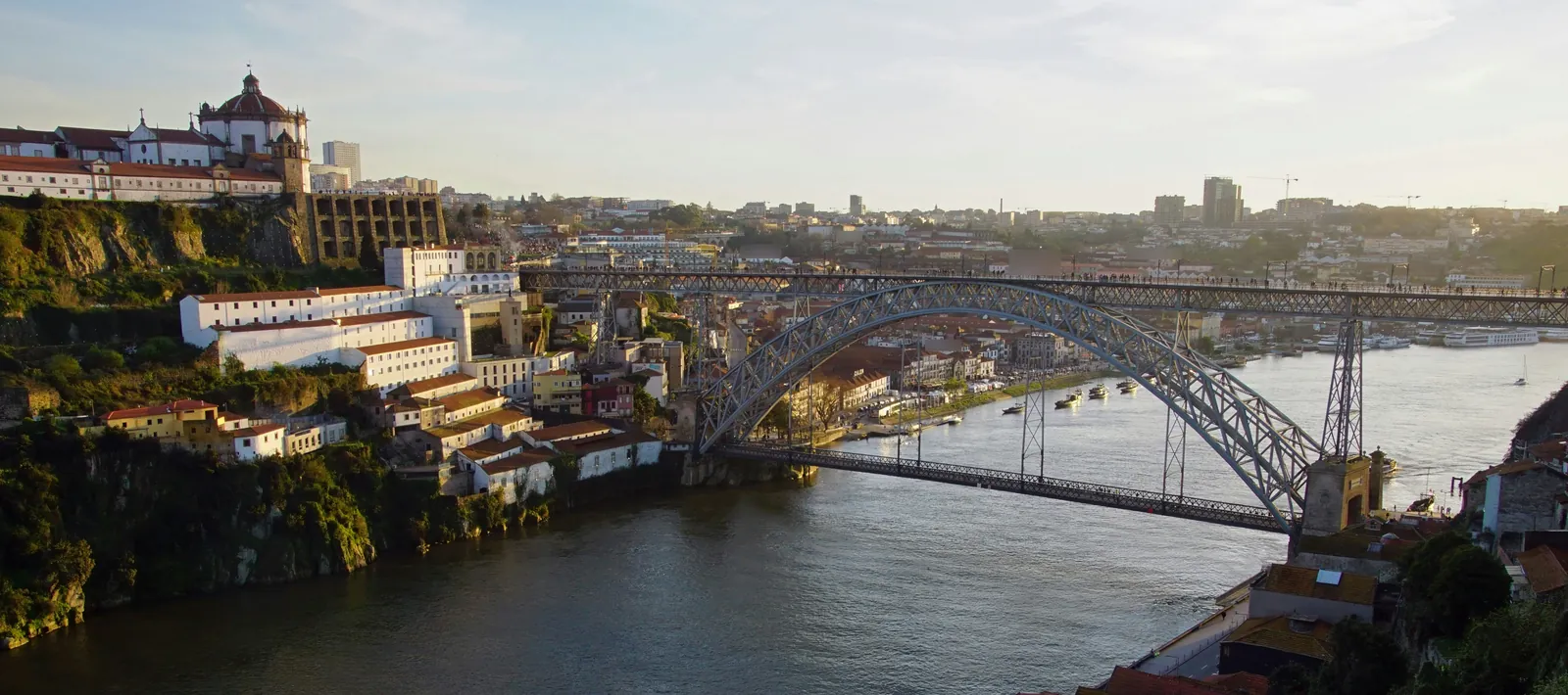 Iron arch bridge over a wide river at dusk, with a domed monastery on the cliff to the left and the city spread beyond