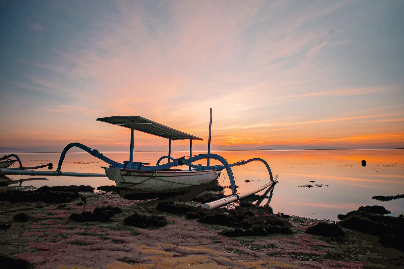 A traditional outrigger fishing boat sits on the reef flat at low tide at sunrise, with a calm pink and orange sea and low hills on the horizon