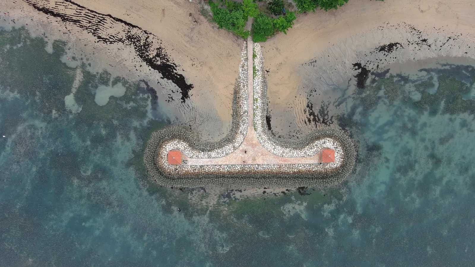 A T-shaped stone breakwater with two small red-roofed pavilions, seen from straight above, with sand, beach trees and green water over the reef