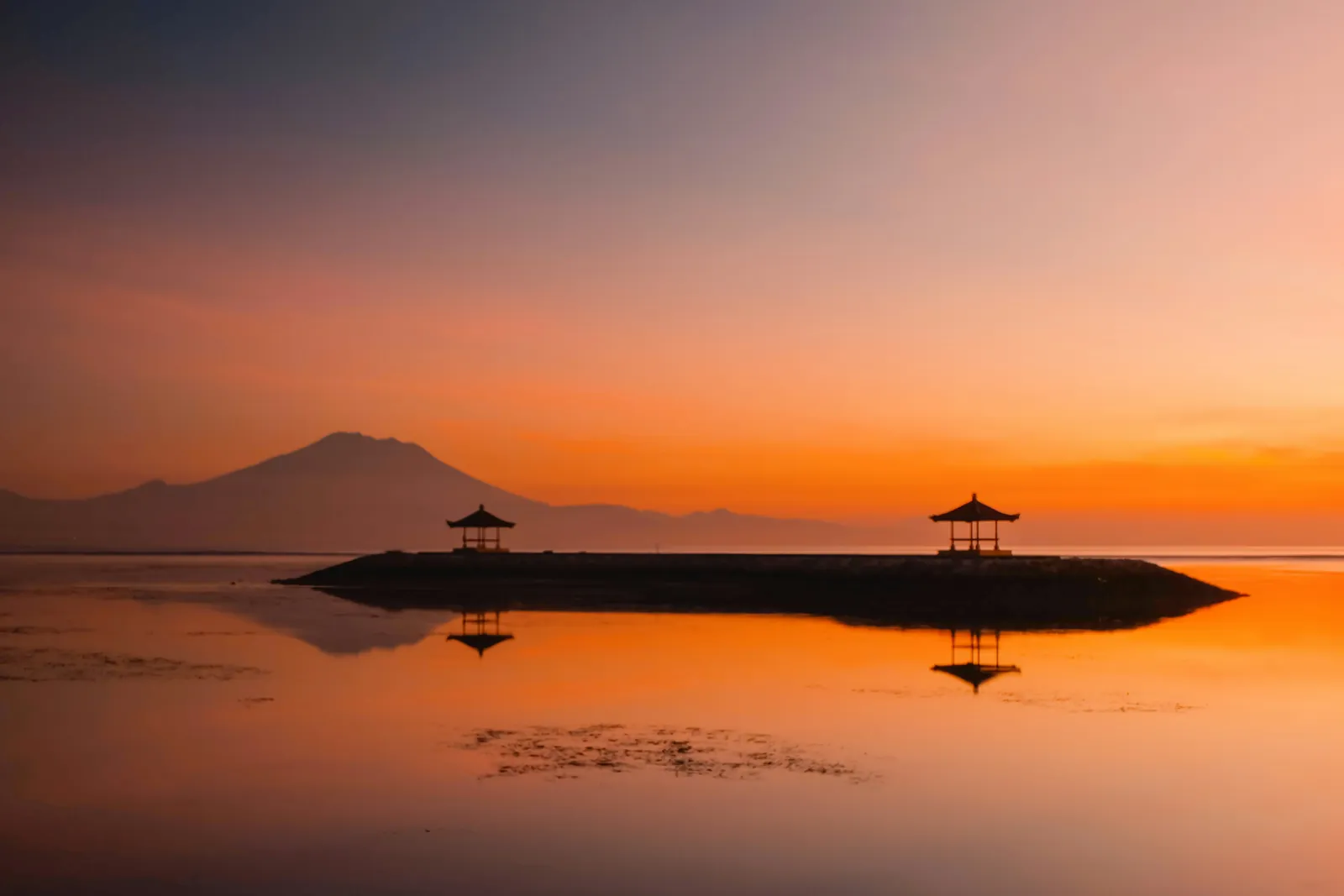 Sunrise over a still sea, with a stone breakwater carrying two small pavilions and a volcano on the horizon, all in orange light reflected in the water