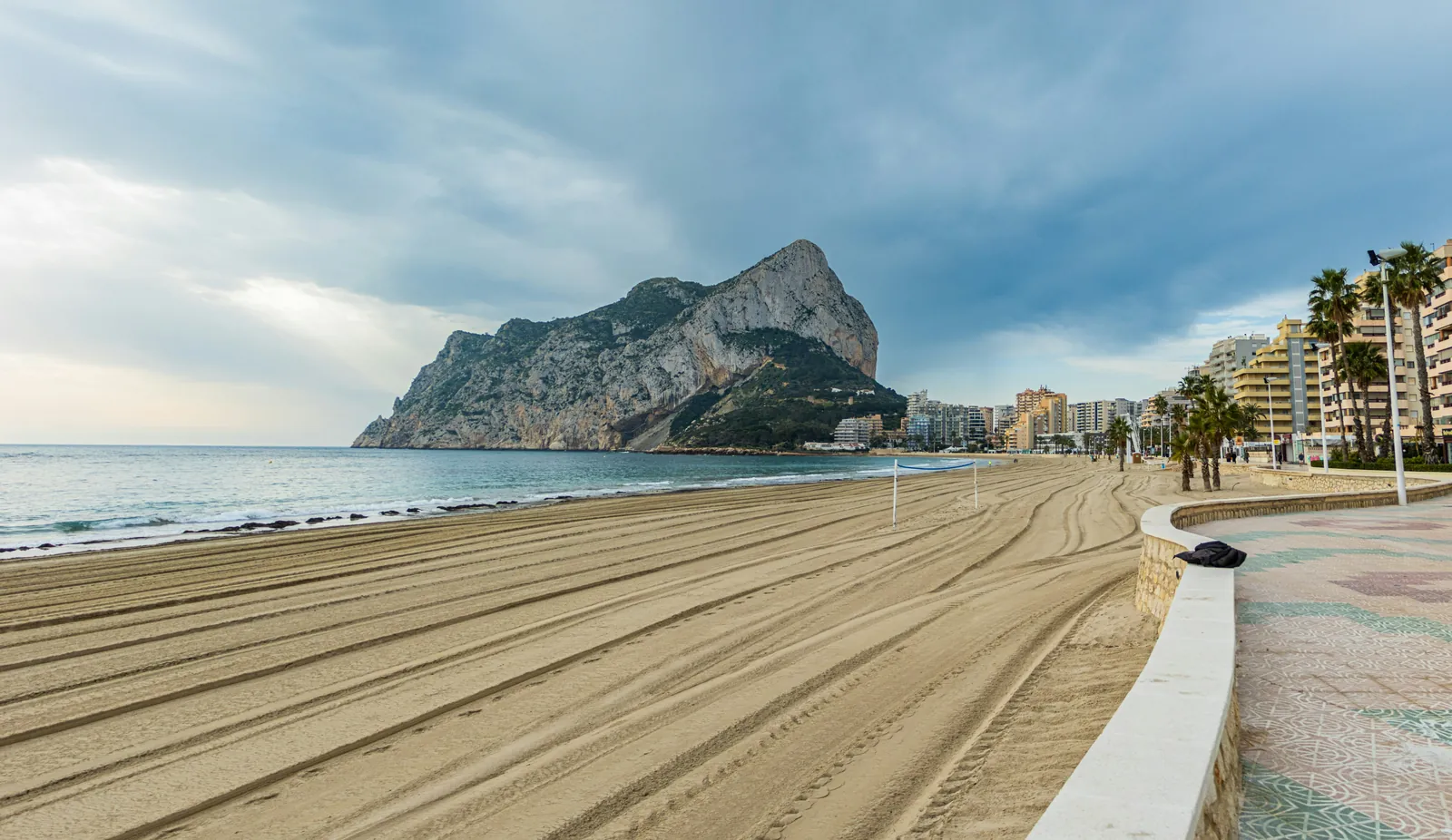 Wide empty beach raked into curving lines, with a tiled promenade on the right, apartment blocks and the huge Peñón de Ifach rock behind
