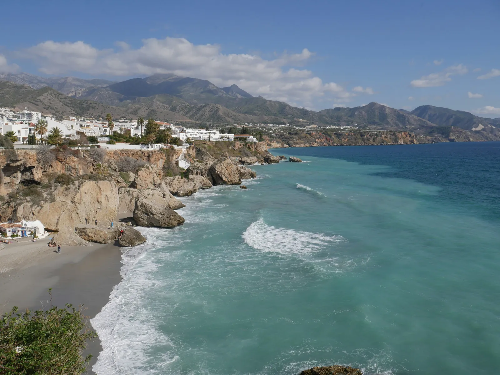 Rocky cliffs and a small sandy cove with surf rolling in, white houses along the clifftop and grey mountains behind