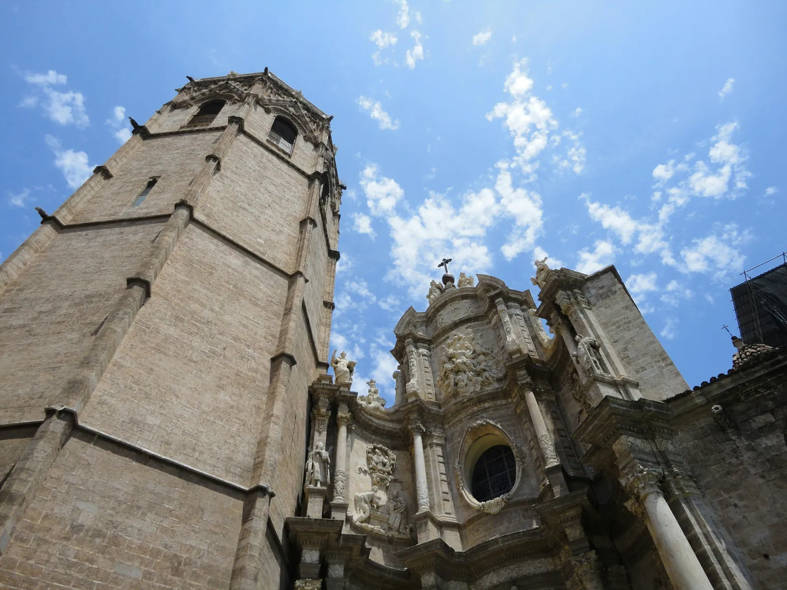 Looking up at a tall octagonal stone bell tower beside an ornate baroque church doorway, against a blue sky with small clouds