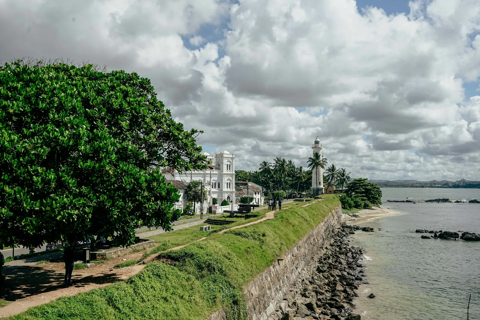 A grassy stone rampart running along the sea towards a white lighthouse among palm trees, with a white colonial-era building and a large shady tree on the left