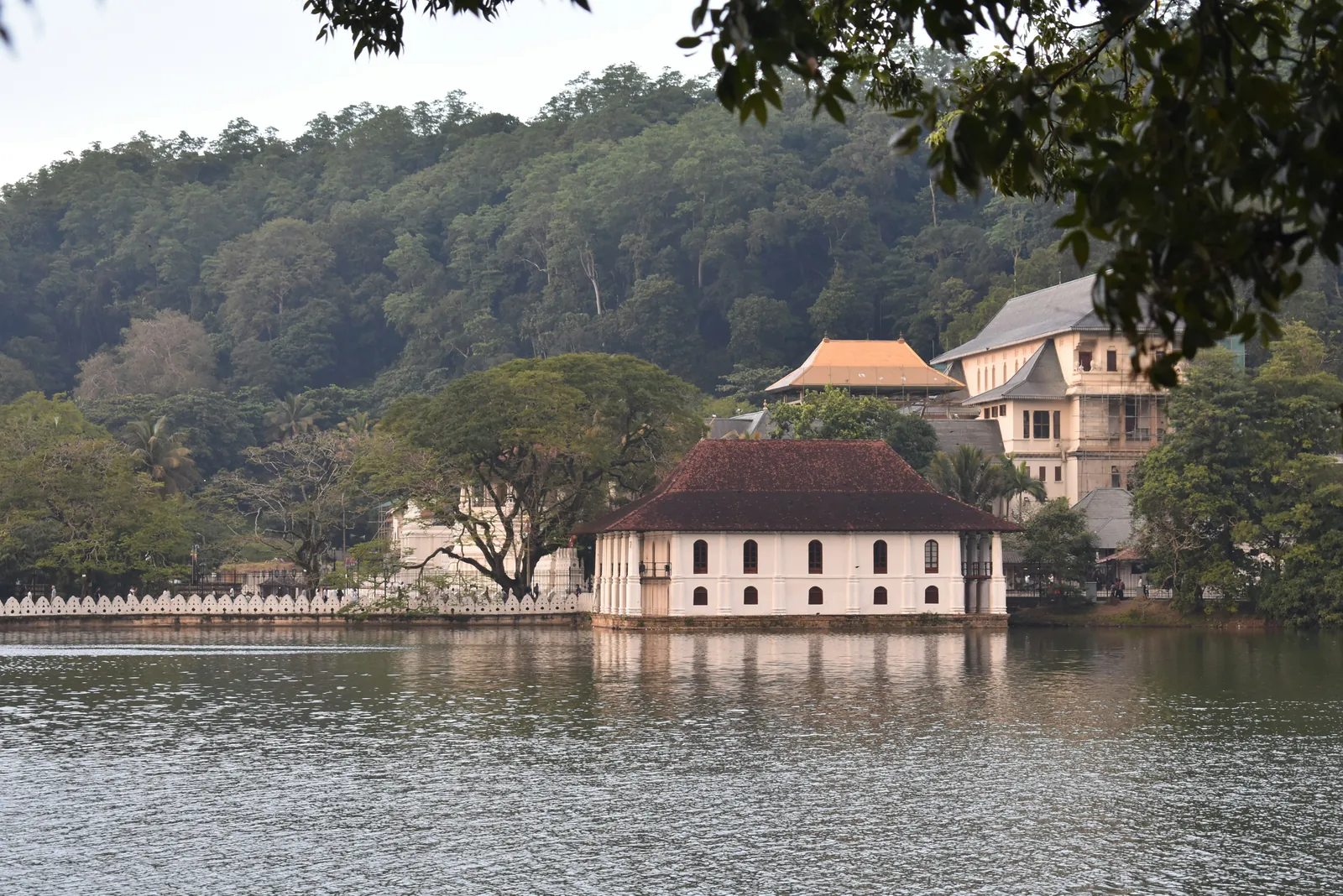 A white pavilion with a dark tiled roof standing at the edge of a calm lake, a scalloped white wall running along the shore and forested hills behind