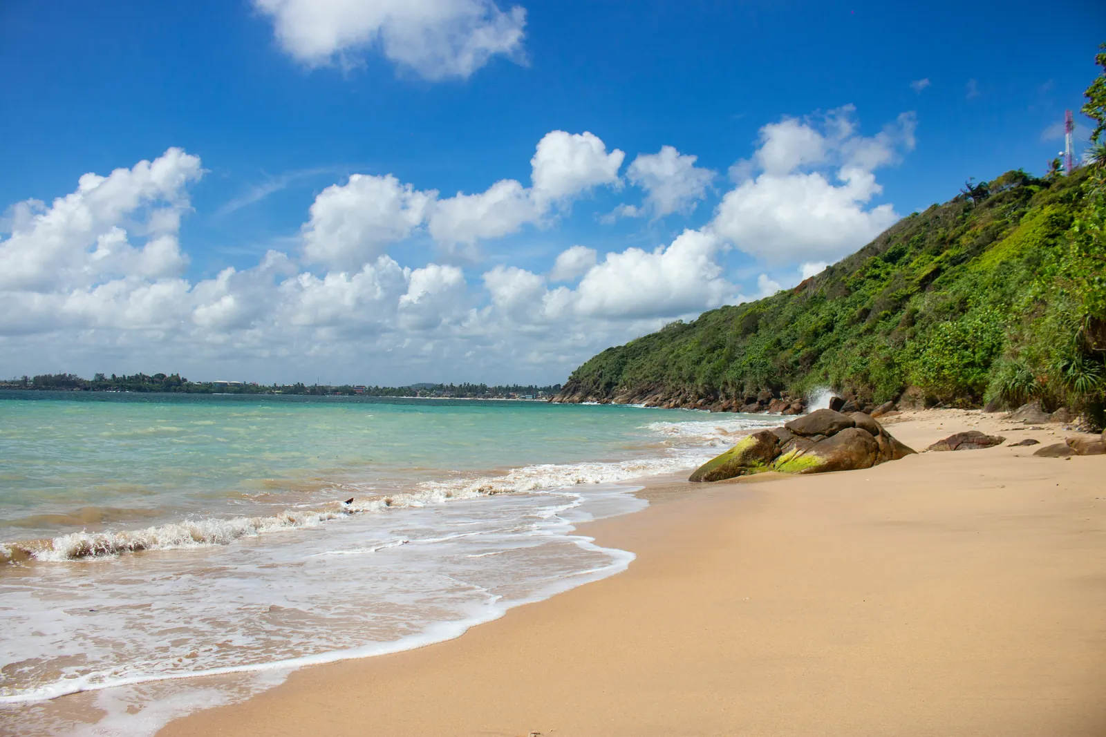 An empty golden sand beach with small waves, a mossy boulder and a steep jungle-covered headland on the right, under a blue sky with white clouds