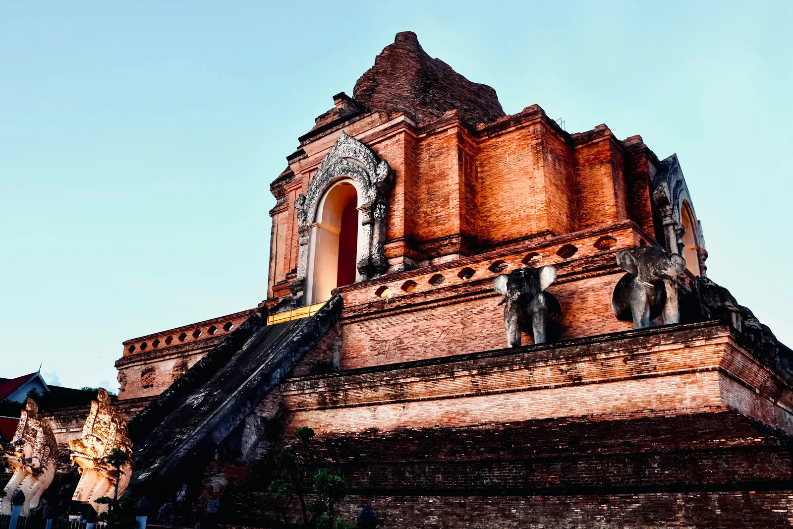 Ruined red brick chedi of Wat Chedi Luang with stone elephants on its ledge and carved naga heads at the foot of the stair, against a pale evening sky