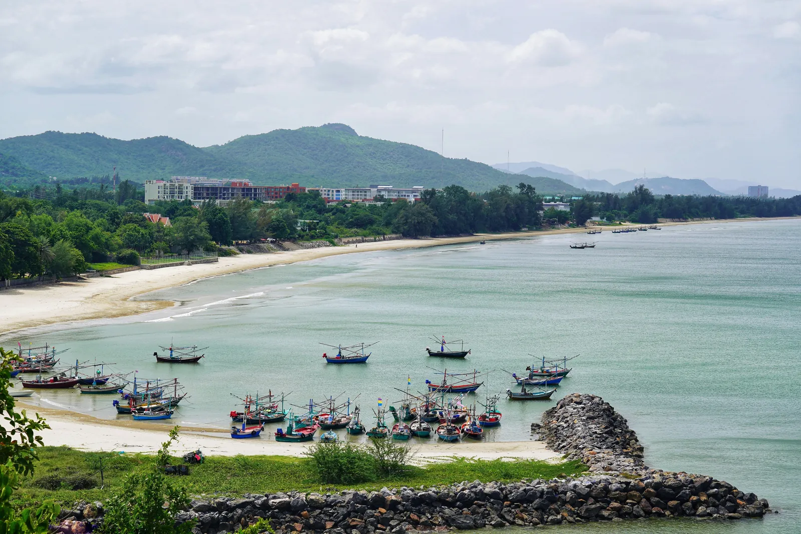 Curving sandy beach with colourful fishing boats moored in a calm bay, a rock breakwater in front and green hills behind